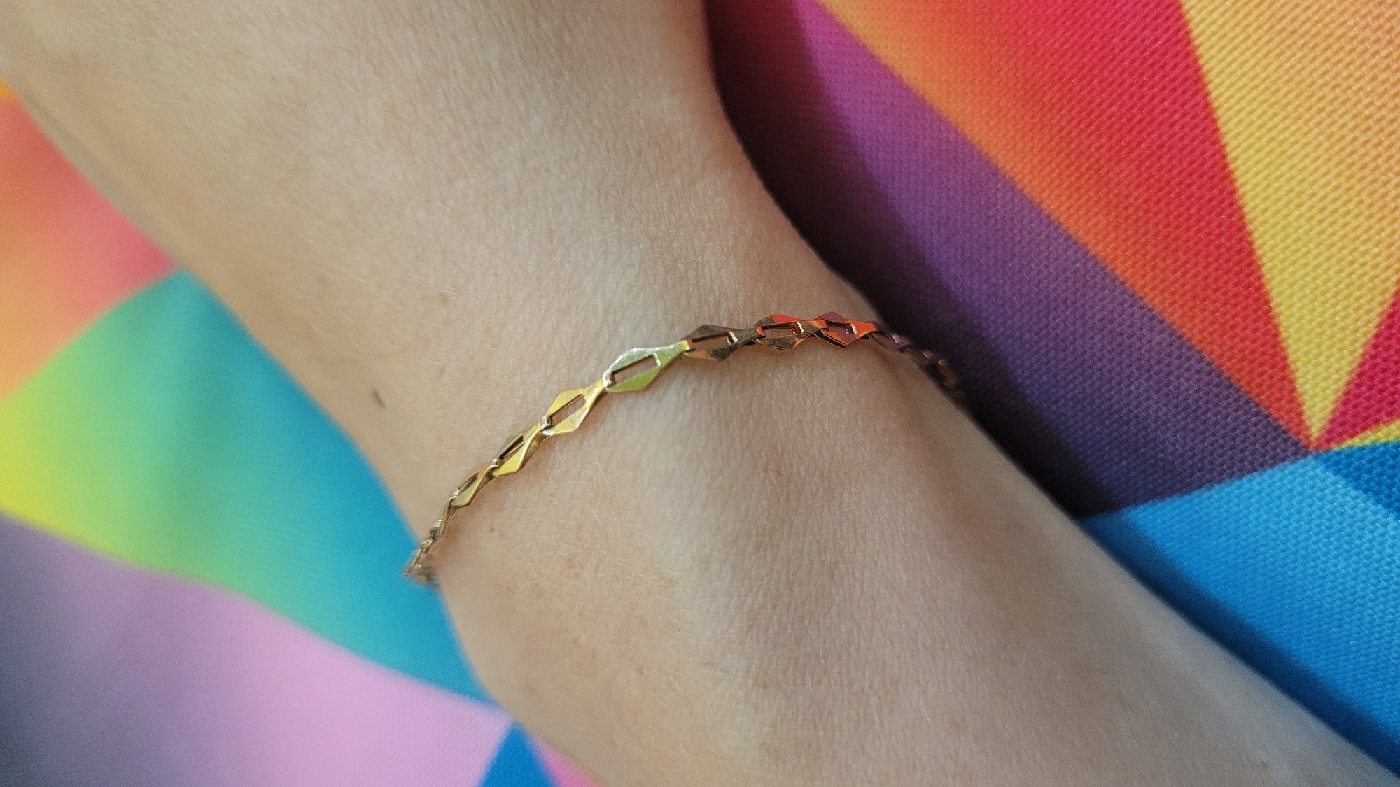 The media file is a close-up shot of a person's wrist and arm, adorned with a delicate gold-colored bracelet. The bracelet features a repeating geometric pattern of interconnected diamond-shaped links. The skin of the arm is fair and appears smooth. The background is a vibrant, out-of-focus pattern of rainbow-colored stripes, radiating outwards like spokes. The colors in the background include shades of blue, purple, red, and yellow, suggesting a fabric or a brightly colored surface. The lighting is bright, highlighting the metallic sheen of the bracelet and the texture of the skin and background fabric. The composition focuses on the jewelry and the arm, with the colorful background providing a stark contrast. No text is visible in the image. The overall impression is one of simple elegance against a bold and cheerful backdrop. The image seems to capture a moment of personal adornment.