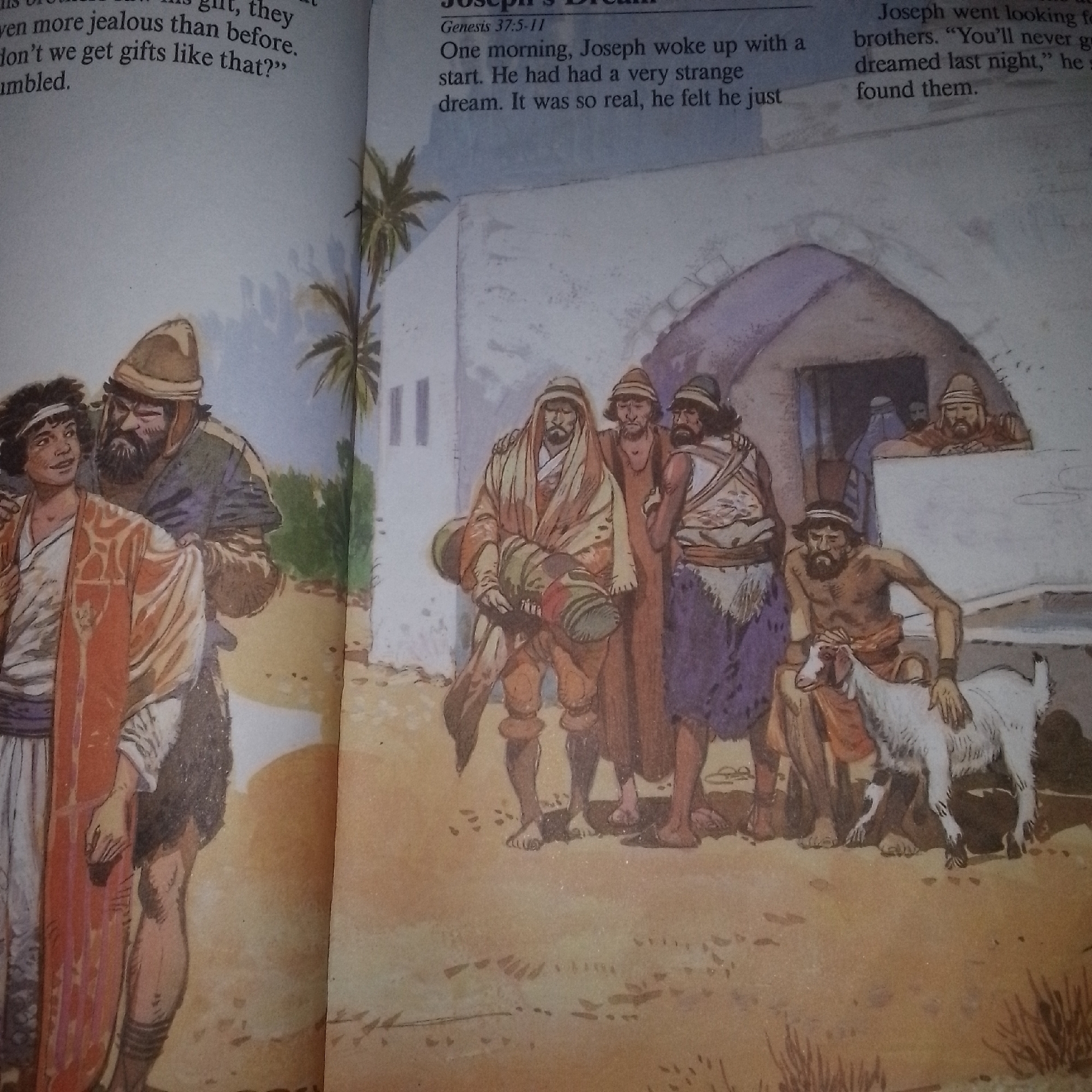 The image depicts a scene from a story, likely biblical, with text visible on the left and right margins. In the foreground, on the left, two men are conversing. One man, younger with dark curly hair, wears a reddish-orange robe over a white tunic. The other, older man, has a dark beard and wears a brown hat and a purple garment. He has his hand on the younger man's shoulder.
The center of the image shows a group of three men walking together. The man in the front carries a large rolled-up object, possibly a mat or fabric, slung over his shoulder. He is dressed in earth-toned robes. The other two men are behind him, one with his arm around the other. They are also dressed in similar ancient attire.
To the right, near a rustic building with a thatched roof, a man is kneeling and petting a white goat. Other figures are visible within or near the building entrance, including two men looking out. The environment appears to be arid, with sandy ground and sparse vegetation, suggesting a location in the Middle East or a similar climate. The overall impression is one of an ancient, rural setting.