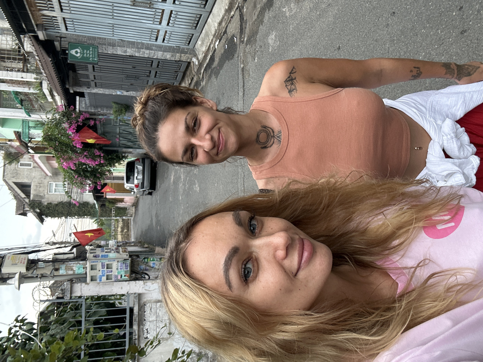 Two women pose for a selfie in a street setting. One woman is positioned in the foreground, with long blonde hair and wearing a light pink top with a pink graphic. Behind her, another woman smiles, sporting a brown tank top and a tattoo on her neck. The scene unfolds in the city of Đồng Nai, Vietnam, with a narrow street lined by buildings, some adorned with Vietnamese flags. A parked car is visible in the background. Above, a gray metal gate and overhanging structures shade part of the street.