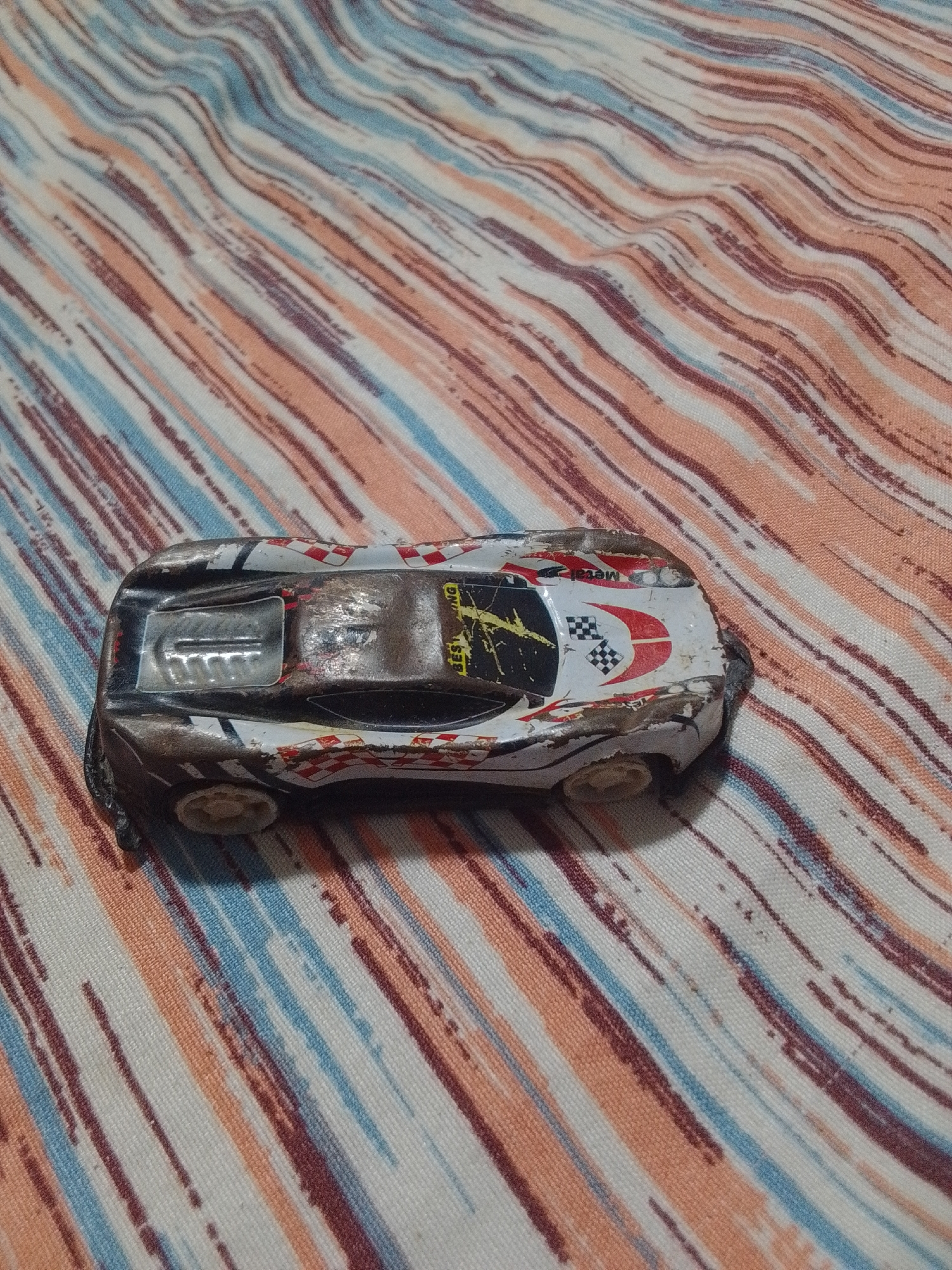 A close-up, slightly elevated shot shows a battered, white toy race car with black and red accents resting on a striped fabric surface. The car appears to be made of metal and plastic, with visible wear and tear, including scratches and chipped paint. The fabric beneath it features diagonal stripes in shades of peach, white, brown, and blue. The lighting is somewhat dim, casting subtle shadows. The image does not depict any people, actions, or interactions, nor does it provide any information about its location in La Tagua, Venezuela.