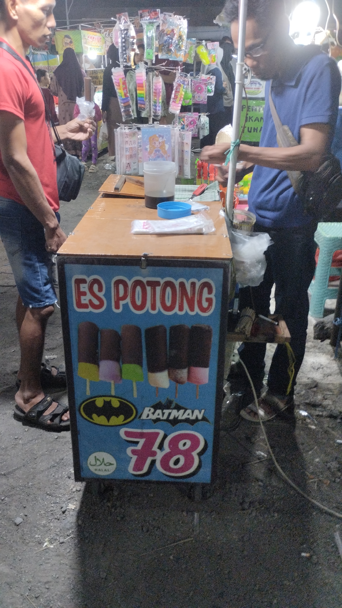 The image depicts a street vendor selling ice cream in Sambungrejo, Indonesia. The central subject is an ice cream cart, a freezer chest with a wooden top, adorned with a vibrant blue graphic advertising "ES POTONG" (cut ice cream) and featuring images of various ice cream bars, the Batman logo, and the number "78". A "HALAL" certification is also visible.
In the foreground, a man wearing a red t-shirt and denim shorts stands on the left, holding a small item in his hand. On the right, another person, wearing a blue t-shirt and dark pants, is partially visible, appearing to interact with the cart or serve a customer. The background reveals a bustling market or roadside stall setting with other vendors and displays of merchandise, including colorful toys and accessories. The lighting suggests it might be evening or night, given the artificial lights illuminating the scene. The overall atmosphere appears lively, characteristic of a local market. Visible text includes "ES POTONG", "HALAL", "BATMAN", "78", and some partial Indonesian text on signage in the background, such as "JASUK MANG: 6, AR: 11", "PENT", "KAN", and "TUN".