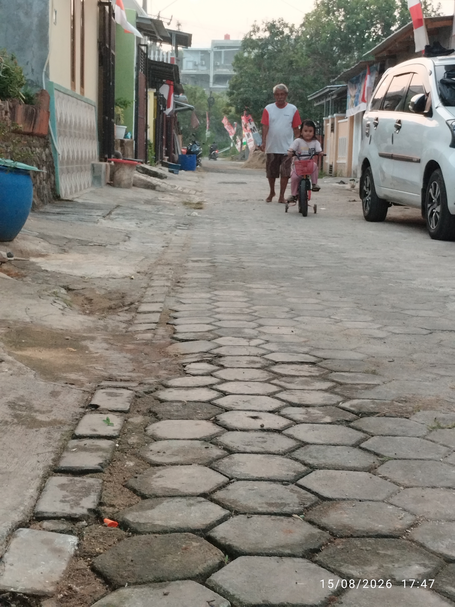 In Bandar Lampung, Indonesia, a street scene unfolds with a young child riding a small tricycle accompanied by an adult. The child, with dark hair, wears a pink shirt and pink pants, her hands gripping the handlebars. The adult, an elderly male with gray hair, is dressed in a white t-shirt with red trim and brown shorts, walking behind the child. To the right of the scene, a white car is parked, its front wheel turned. The street is paved with hexagonal concrete blocks, and a portion of a blue cylindrical object is visible on the left side of the frame, possibly a large water container. The background shows houses with red and white Indonesian flags displayed, indicating a national holiday or celebration. The overall lighting suggests it is late afternoon or early evening.
