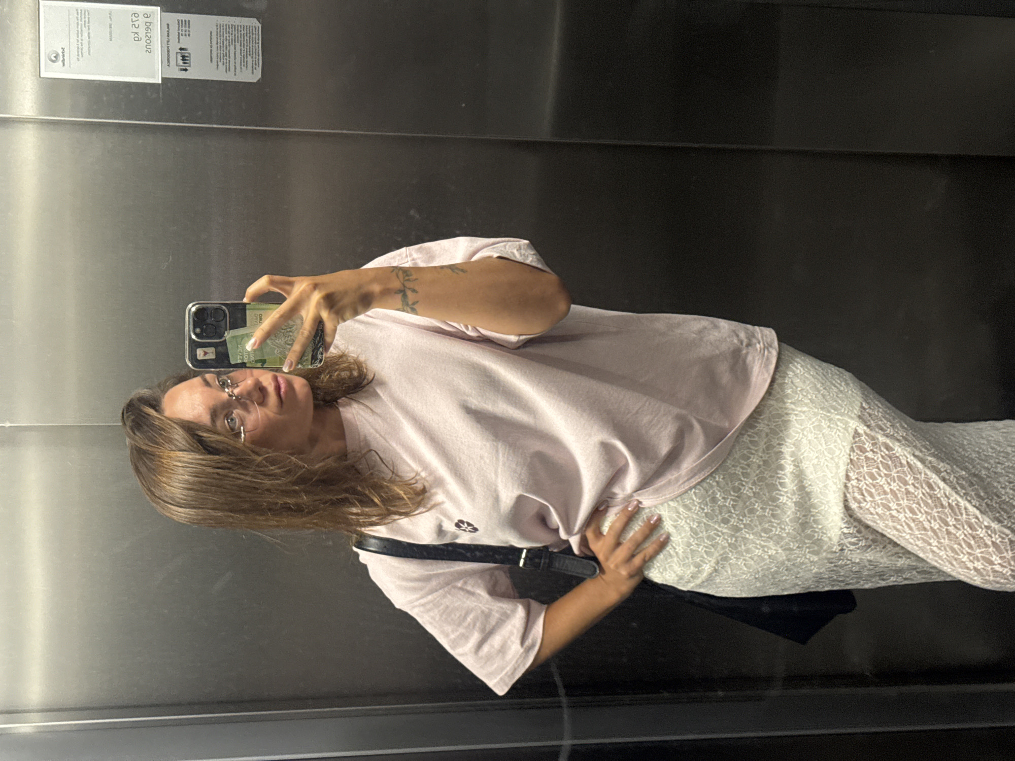 A woman is captured in a mirror selfie, standing in what appears to be an elevator or a room with brushed metal walls. She holds a dark-cased smartphone in her right hand, positioned to take the photo, while her left hand rests on her hip. She has medium-length brown hair, wears clear-rimmed glasses, and has a neutral expression.
Her attire consists of a light pink, short-sleeved t-shirt with a small dark emblem on the left chest, and a white, lace overlay midi skirt. A black shoulder bag with a silver buckle is slung over her left shoulder, visible on her right side. A leafy vine tattoo is visible on her left forearm, and her fingernails are painted a light pink.
The metallic walls of the setting are reflective, showing reversed text from a sticker or plaque. Visible text includes "200.000m" (likely a reflection of "m00.000.002"), "0,2 yd" (from "b.y 2,0"), "380 000W", "www.vll.co.vu", and "zolbotoa" (from "aoboloz"). Tucked into the back of her phone case is a green bill or card, partially showing "BOLIVARES" and "CIEN," indicating a 100 Bolívares note. The scene suggests an indoor public or semi-public space in Budva, Montenegro.