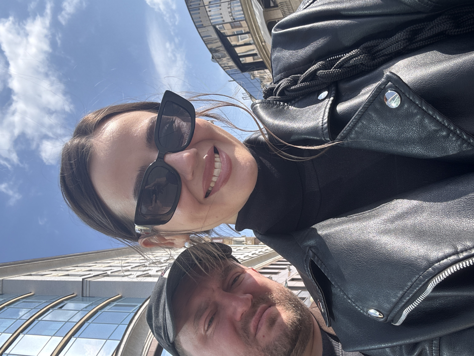 A selfie shows a woman and a man in Moscow, Russia. The woman is in the upper part of the frame, smiling with sunglasses and wearing a black turtleneck and a black leather jacket. The man is in the lower part of the frame, wearing a cap and a shirt. Behind them, buildings reflect in a mirror-like structure. The sky is blue with light clouds.