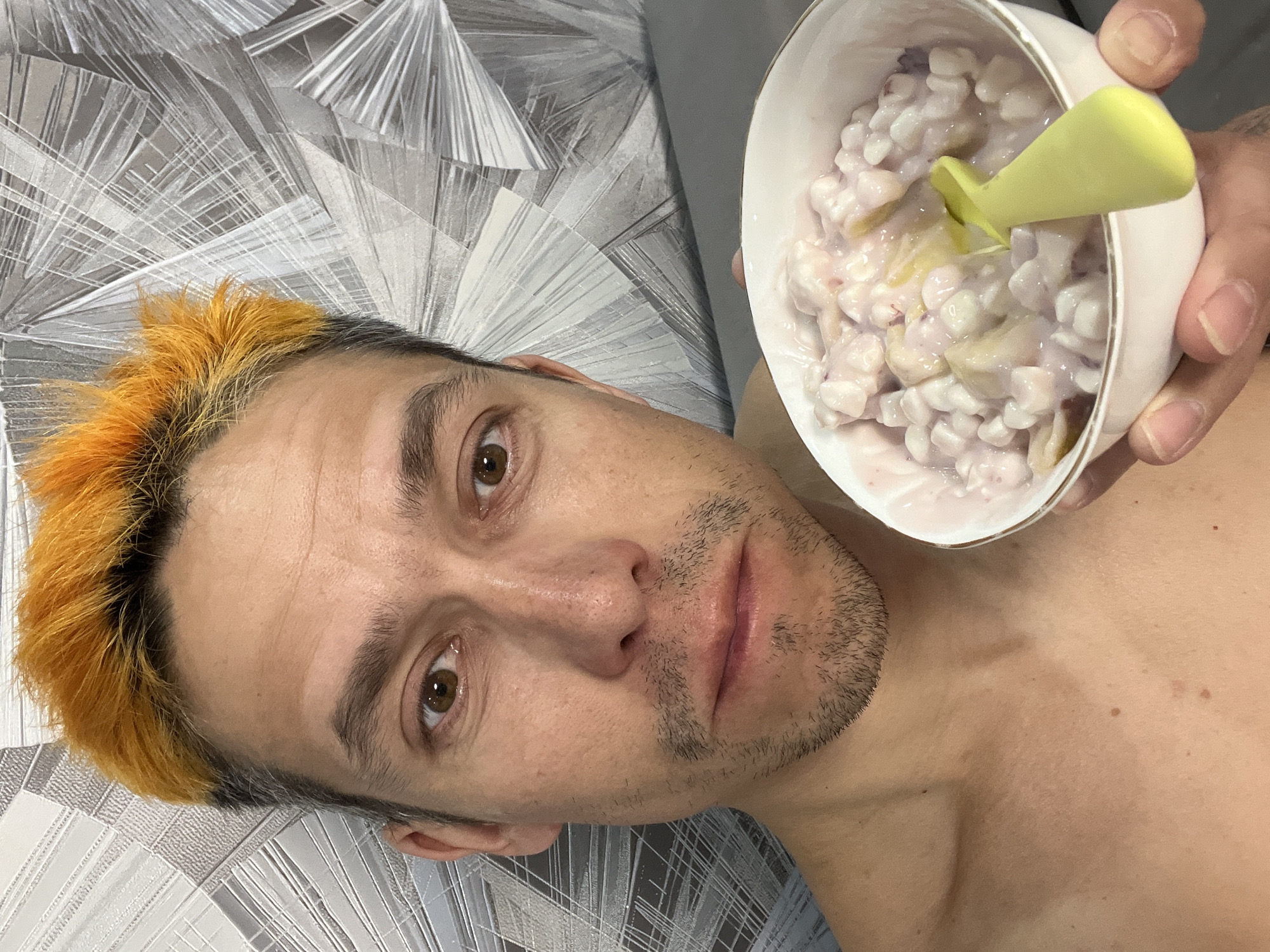 A selfie of a man is taken indoors. The man has short, dyed orange hair on top and is topless. He holds a white bowl of food with a green spoon in his right hand. The bowl appears to contain a mixture of white and light purple components, potentially yogurt-based. The background appears to be a gray, patterned surface, possibly bedding or upholstery. No specific details suggest the location is within the city of Chaikovsky, Russia, as that information cannot be derived from this image.
