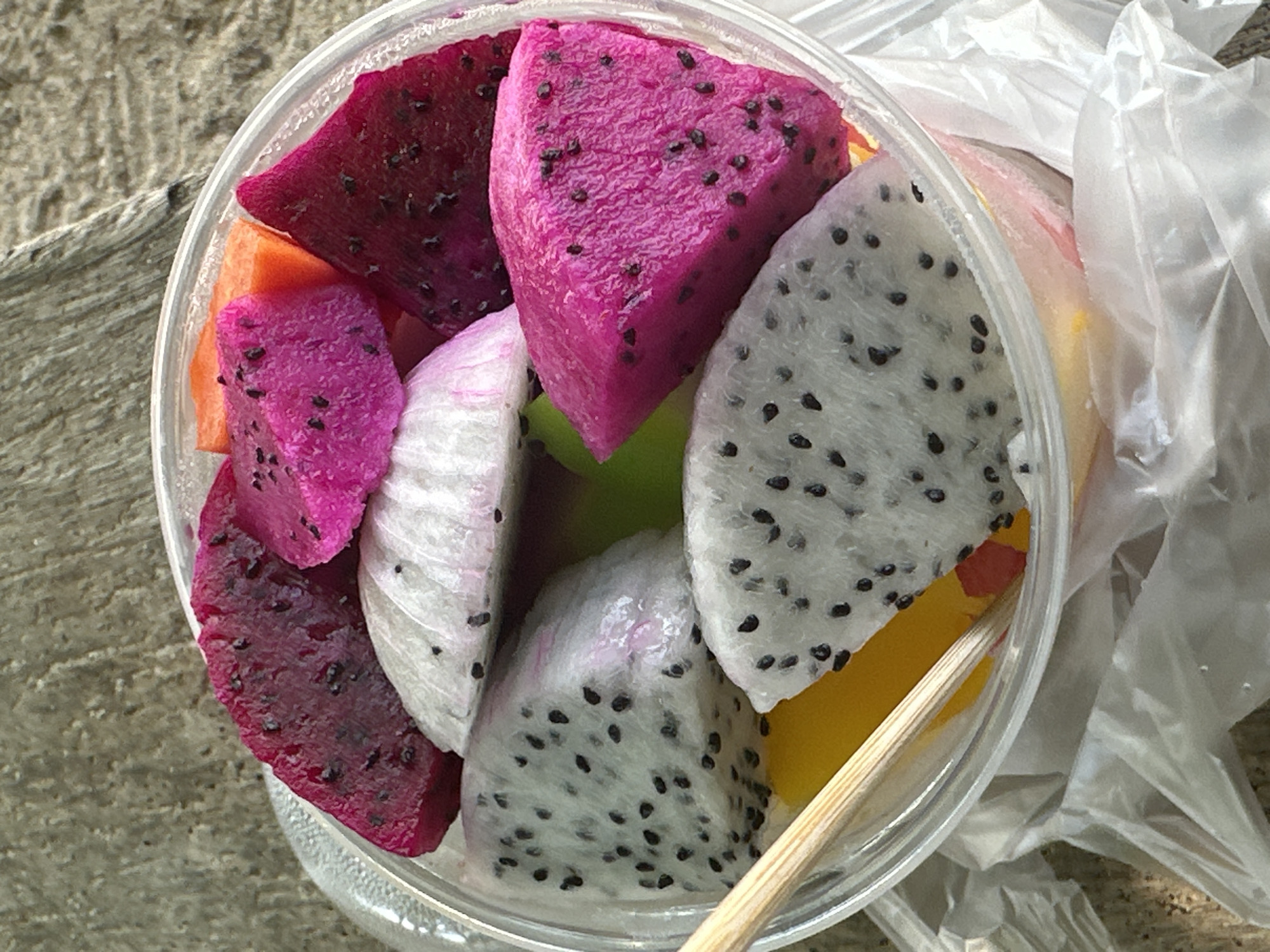 A clear plastic cup filled with sliced tropical fruits, including white and pink dragon fruit and some orange fruit, sits on a rough, textured surface. The cup is partially obscured by white plastic packaging. A wooden skewer is inserted into the fruit. The overall setting suggests an outdoor scene, possibly a market or street food vendor.