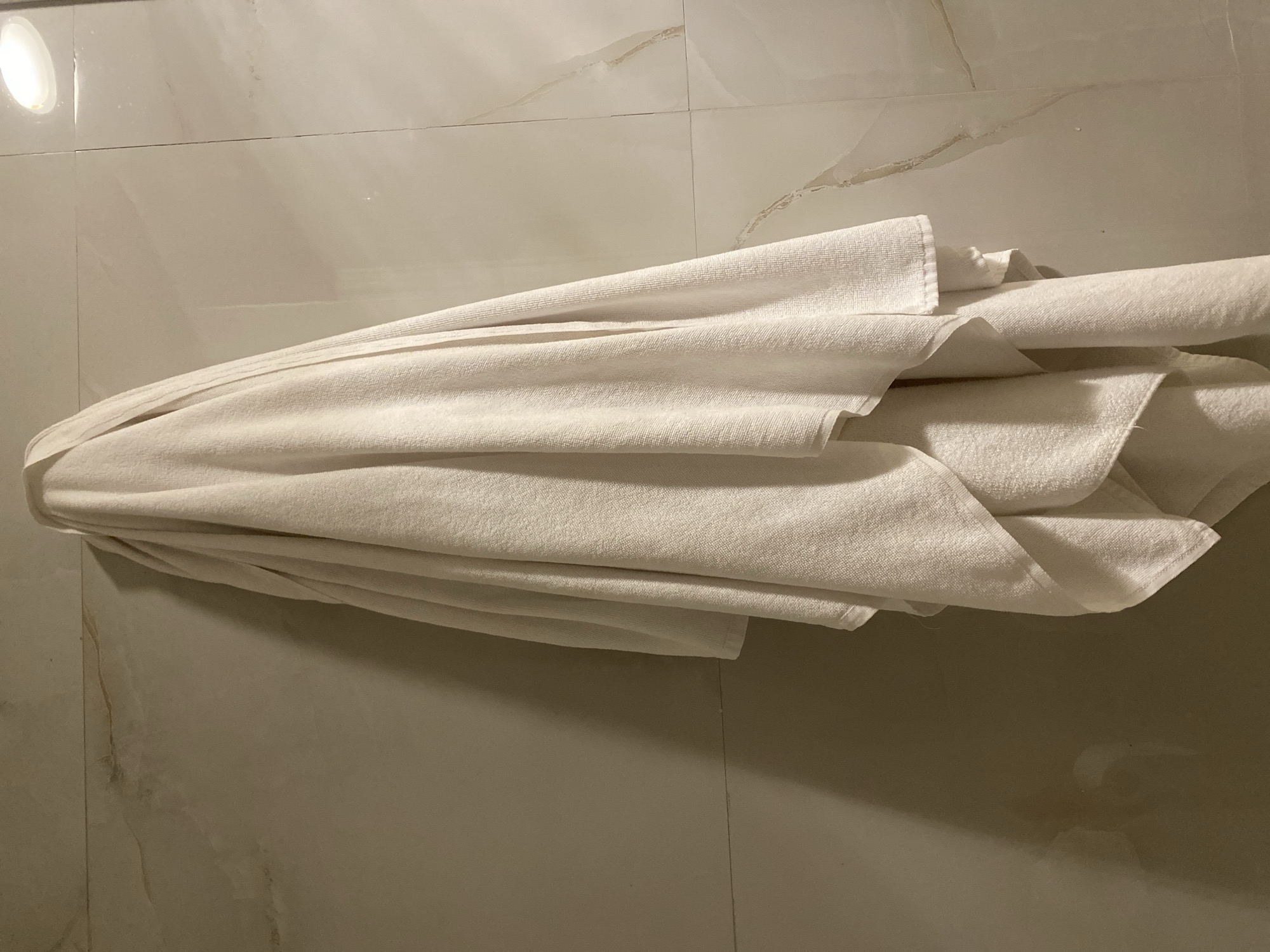A white towel is draped over a hook against a tiled wall. The wall appears to be made of large, light-colored tiles with subtle veining. The towel is thick and textured, with visible folds and creases. The lighting suggests an indoor environment, possibly a bathroom or a changing room. There are no people or animals visible in the image. The scene is static and unremarkable.