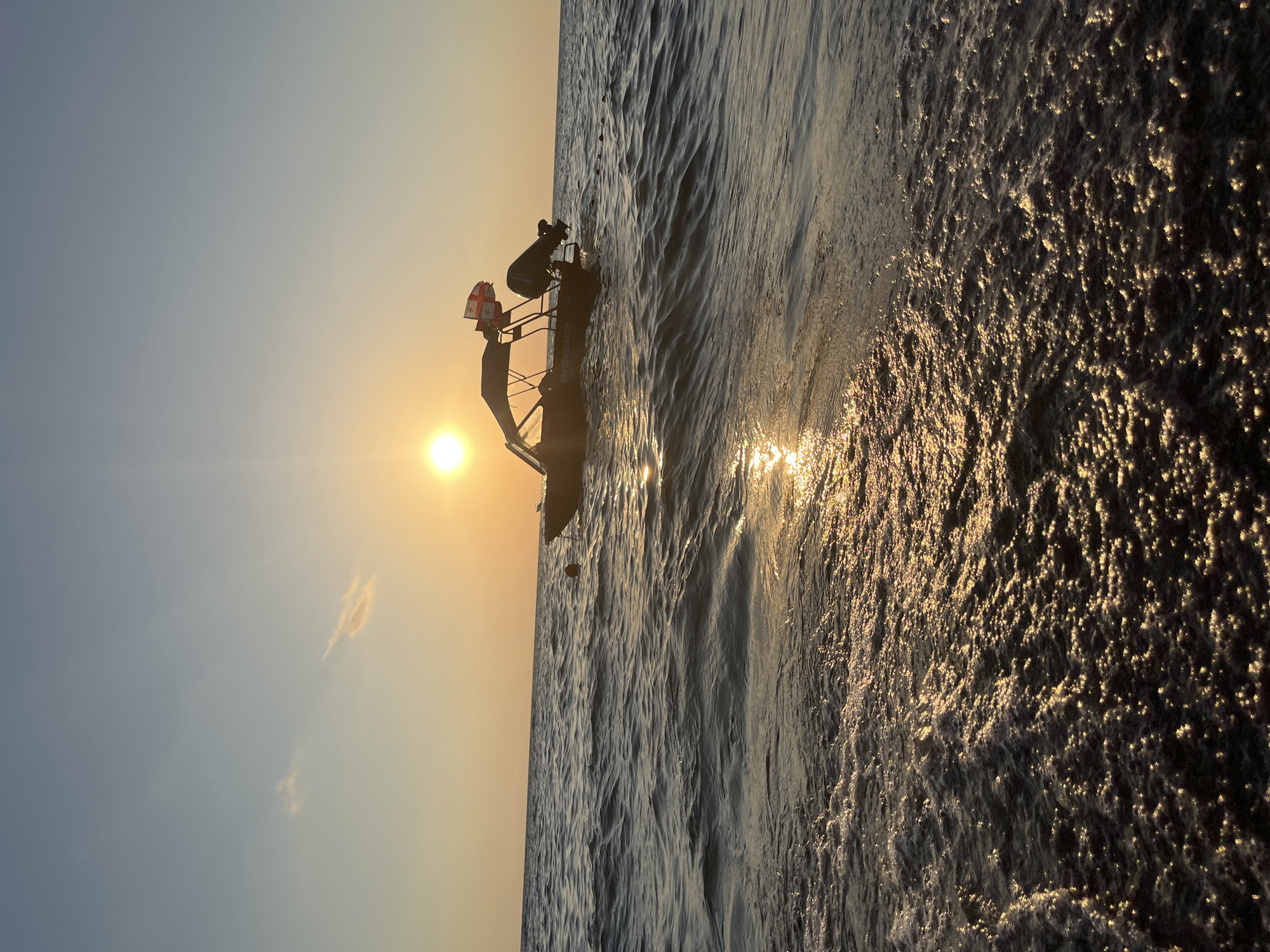 An outdoor, eye-level shot captures a boat on the Black Sea near Ureki, Georgia, at sunset. The sun, a bright circle, hangs low in the hazy sky, casting a golden glow on the water. The boat, dark and equipped with an outboard motor and two flags (one appearing to be the flag of Georgia), is anchored in the choppy, dark water. The wake of the boat is visible in the foreground, a frothy white trail against the darker sea. The horizon is clearly defined, separating the water from the pale orange and blue gradient of the sky, where a single wispy cloud drifts. The overall scene evokes a sense of tranquility and the end of a day.