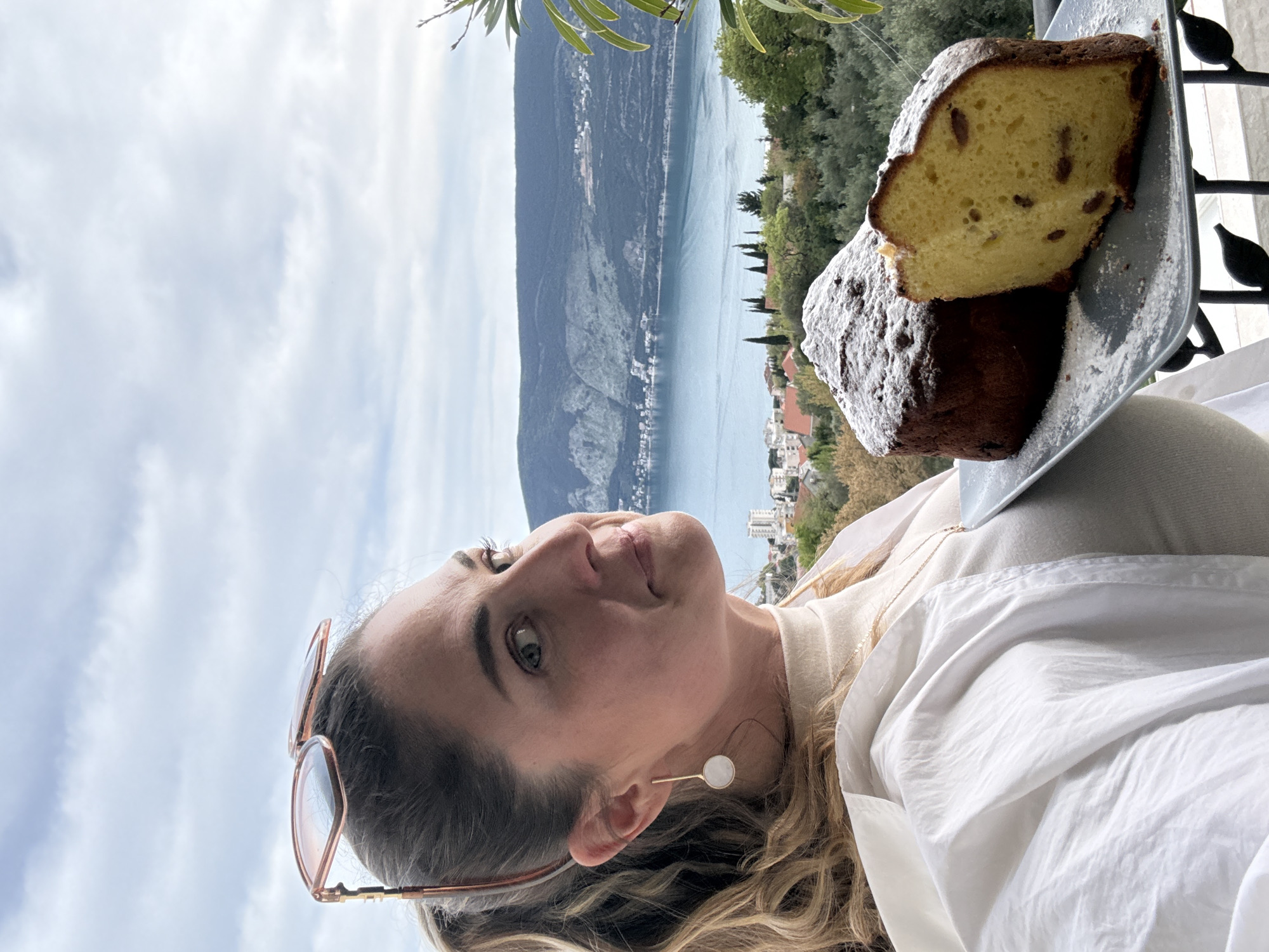 The image is an eye-level portrait of a woman holding a plate of cake against a background of a coastal landscape. The woman has light skin, light brown hair, and green eyes. She wears a white shirt, a beige sweater, sunglasses on her hair, and an earring. On the plate, there are two slices of cake covered in powdered sugar. The background features a body of water, possibly a lake or a bay, with mountains visible on the opposite shore. Trees and buildings can be seen along the coastline. The image does not provide enough information to determine the specific location in Trebesin, Montenegro, but it shows a scenic view typical of coastal regions.