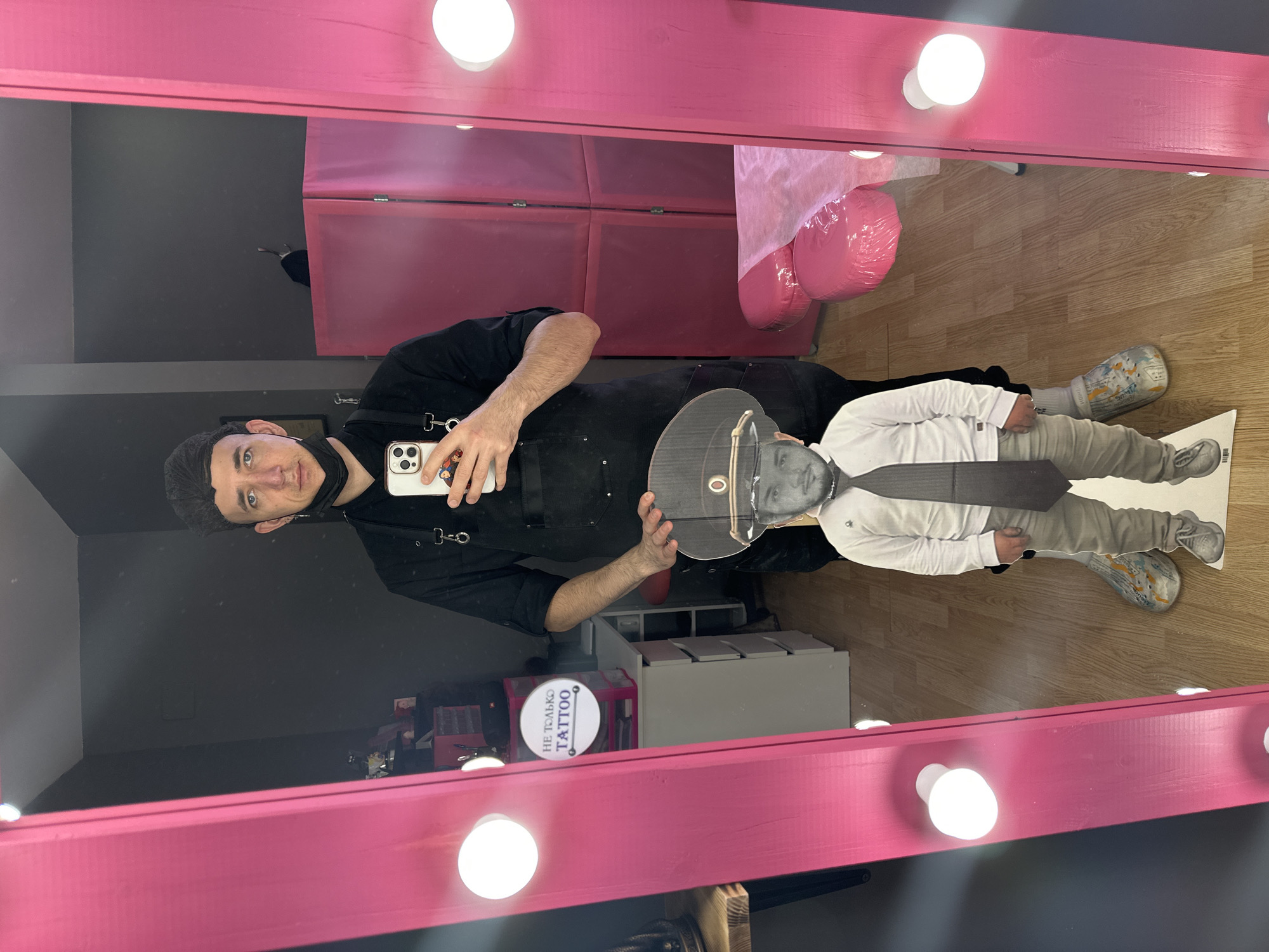 The scene is an interior view of a commercial establishment in Saratov, Russia, identified as a tattoo studio by a visible sign.
A male individual is reflected in a large, vertical, bright pink vanity mirror, which is illuminated by multiple exposed bulb lights along its frame. The man wears a dark, short-sleeved top or apron with chest straps, a black face mask positioned below his chin, and a dark hairnet or cap. He holds a white smartphone, its screen facing the viewer, as if capturing his reflection.
Positioned on the floor directly in front of the reflected man is a cardboard cutout of a small person. This cutout depicts a figure wearing a white long-sleeved shirt, a dark tie, light-colored pants, and a military-style peaked cap.
The background, visible through the mirror's reflection, consists of dark grey walls and a light-colored wood laminate floor. To the left, a white desk with multiple drawers holds various items, including a black portable speaker. Above the desk, a round white sign with the Russian text "НЕ ТОЛЬКО TATTOO" (NOT ONLY TATTOO) is displayed. To the right, a pink folding room divider stands behind a pink upholstered examination or procedure table, which is covered with a clear protective plastic sheet.