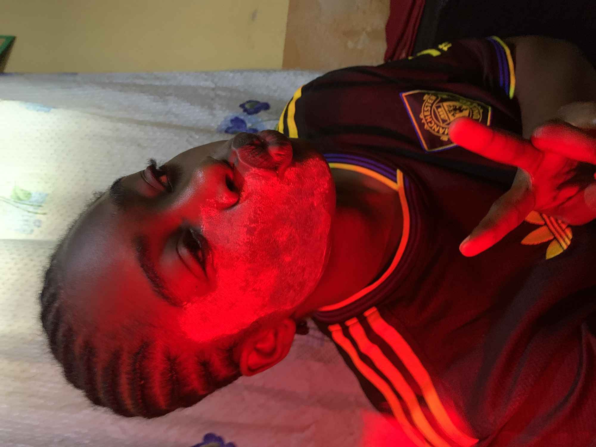 A young Black individual is shown from the chest up, leaning towards the camera with their head tilted. They are wearing a dark Manchester United soccer jersey with yellow and blue trim. Their face is partially obscured by a red light source, giving their skin a reddish hue. They have dark braids and are making a duck face with their lips puckered. Their right hand is raised, showing a peace sign. The background consists of a light-colored curtain with a blue floral pattern and a yellow wall. The image appears to be taken indoors in a room. The location is specified as Rafin Par, Nigeria.