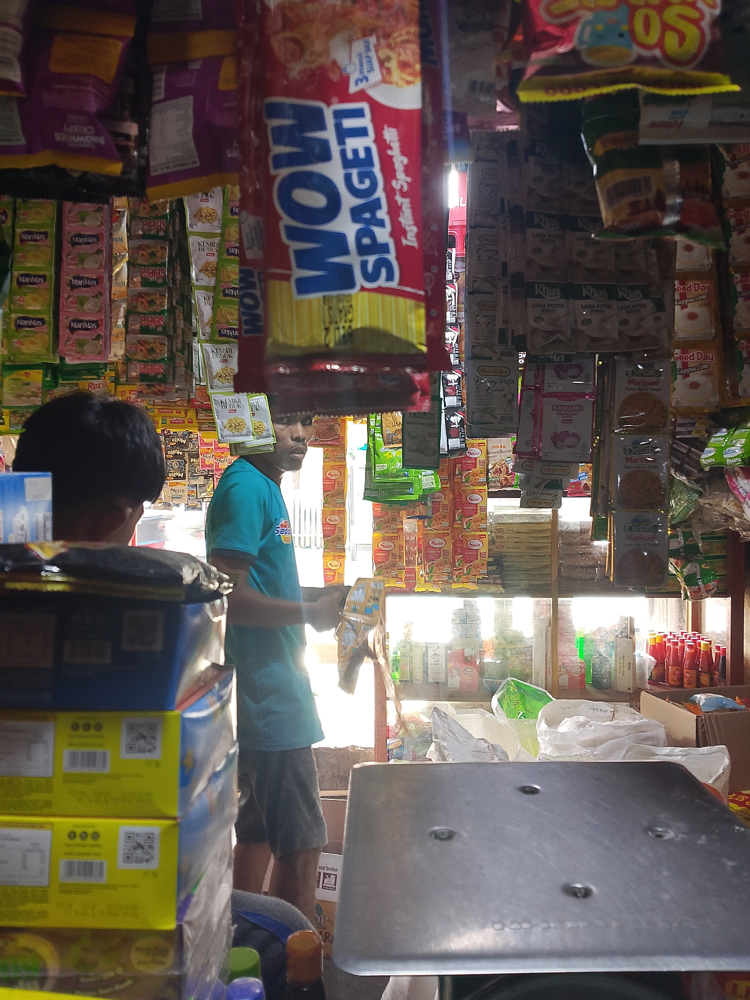 This is a medium shot of the interior of a small grocery store in Palembang, Indonesia. In the foreground, a stack of yellow and blue boxes is on the left. The boxes have QR codes and text that are too blurry to read. To the right of the boxes, a person with dark hair is partially visible, facing away from the camera. In the mid-ground, a man in a teal t-shirt and dark shorts is standing behind a counter, looking towards the right. He appears to be interacting with a customer who is out of frame or obscured. The shelves behind the man are packed with various packaged food items, including bags of spaghetti, instant noodles, and small packets of spices or seasonings. A large red and yellow "WOW SPAGETI" package is prominently displayed hanging from the ceiling. The store appears to be well-stocked but disorganized, with items packed closely together. The lighting suggests it is daytime, with sunlight entering from the right side of the store. The overall impression is of a busy, local convenience store.