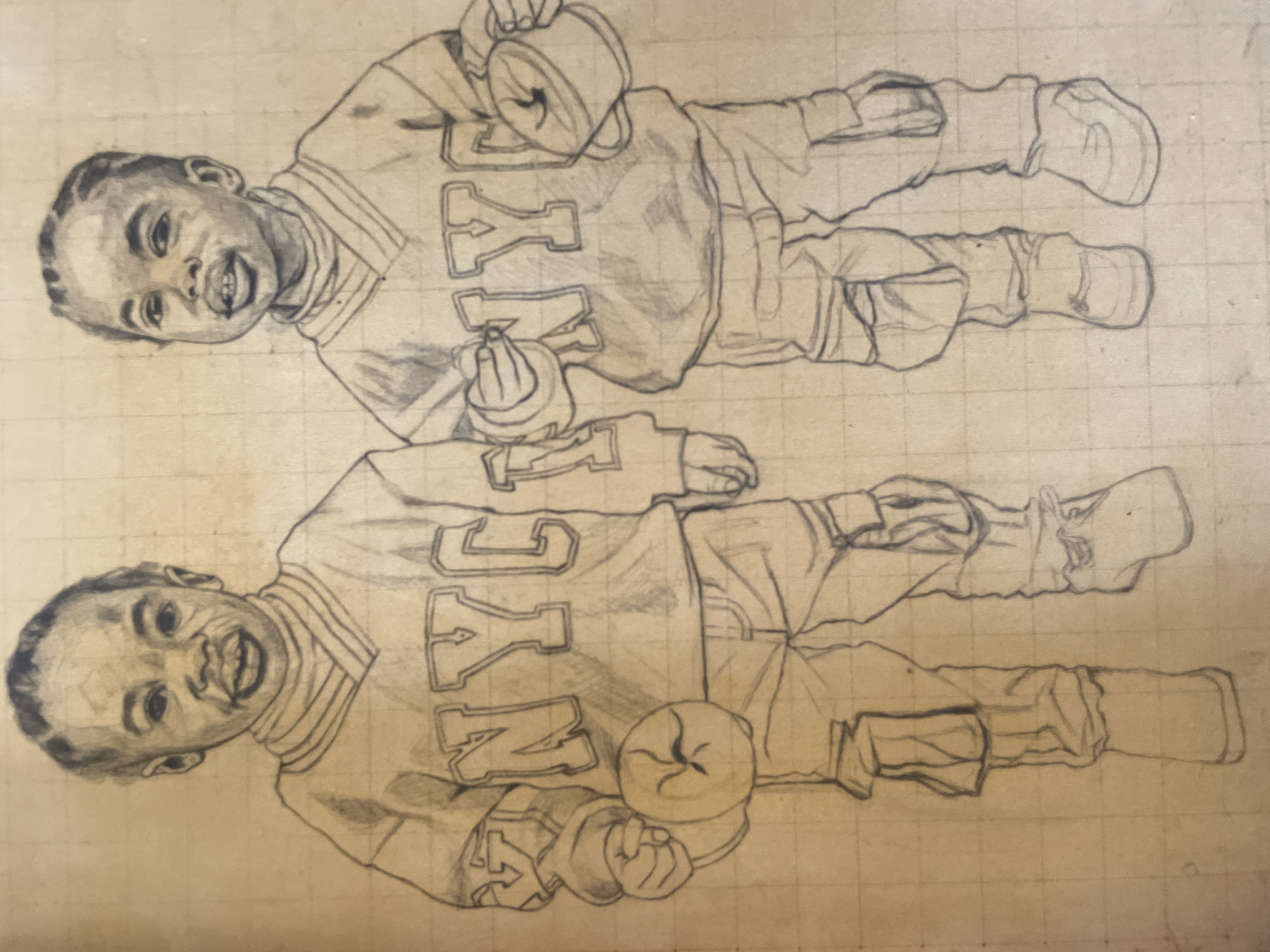 The image shows a pencil drawing of two identical children wearing matching outfits with the word "Kentucky" prominently displayed. The children are both smiling, and they are holding objects: the upper child holds a cylindrical object, while the lower child holds a rounded object. The drawing is on a grid-patterned surface, suggesting it was a sketch or practice piece. There is no evidence to connect the drawing to Ado, Nigeria.