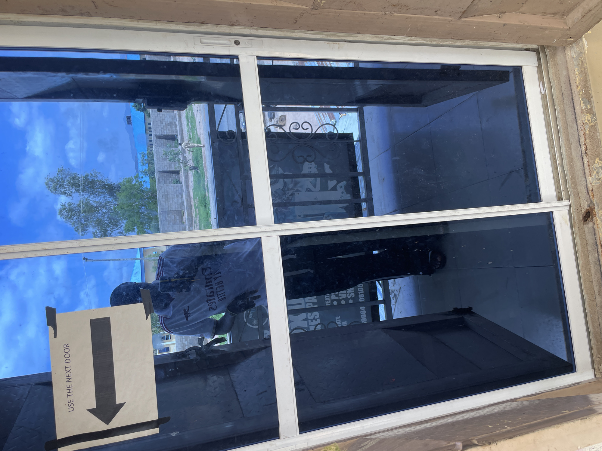 A reflection in a blue-tinted glass door shows a person wearing a white polo shirt with red and white trim, possibly a sports jersey, and dark gloves. A sign taped to the glass reads "USE THE NEXT DOOR" with a black arrow pointing left. Behind the reflective surface, a brick wall and a blue-roofed building are visible, along with trees and mountains in the distance. The scene appears to be outdoors, in a city setting. Based on the details and context provided in other associated images, this location is in Jalingo, Nigeria.