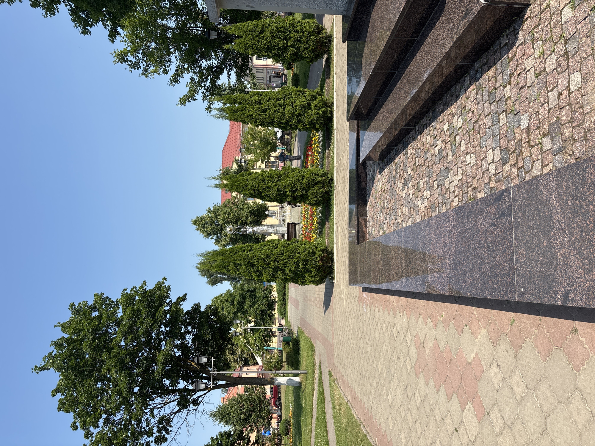 The image depicts a sunlit outdoor scene in a city park. In the foreground, there is a paved area with dark granite steps and a reflective surface. This area transitions to a pathway made of interlocking cobblestones and pavers in shades of beige and pink. To the left, a grassy area with a pathway and a lamppost is visible, leading towards buildings in the background.
Dominating the mid-ground are several tall, conical evergreen trees planted in a line, with a flower bed of red, yellow, and pink flowers behind them. A statue or monument, partially obscured by the trees, stands in the background, along with benches and a fountain.
Further in the background, there are low-rise buildings with red and yellow facades, characteristic of urban architecture. A red car is parked near one of the buildings. The sky is clear and blue.
The image captures a tranquil public space, likely a square or park in the city of Pastavy, Belarus, given the context provided. The scene is orderly and well-maintained, suggesting a public area for recreation or remembrance. The presence of trees, flowers, paved walkways, and buildings indicates a typical urban park or town square. There are a few individuals visible in the background, appearing to be pedestrians. The overall impression is one of a pleasant summer day in a European town.