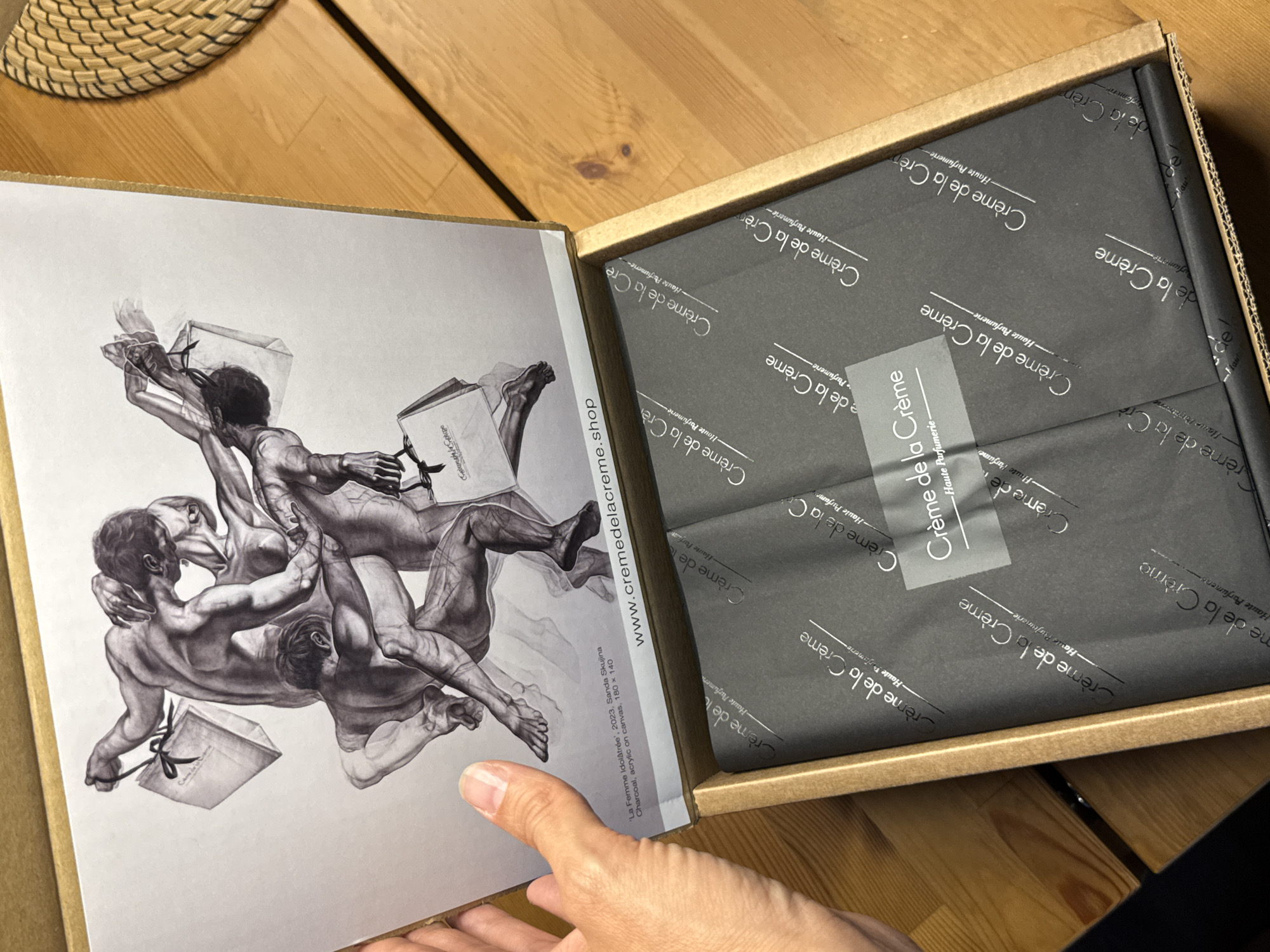 A person's hand is visible in the lower left of the frame, holding open a cardboard box. Inside the box, black tissue paper with a repeating "Crème de la Crème - Haute Parfumerie" logo is visible. The lid of the box contains a print of a charcoal drawing depicting several nude figures intertwined, some holding shopping bags. Text below the print reads "'La Femme Idolâtrée', 2023, Sanda Skujina Charcoal, acrylic on canvas, 180 x 140 www.cremedelacreme.shop". The scene is set on a wooden surface. The location is not specified in the image.
