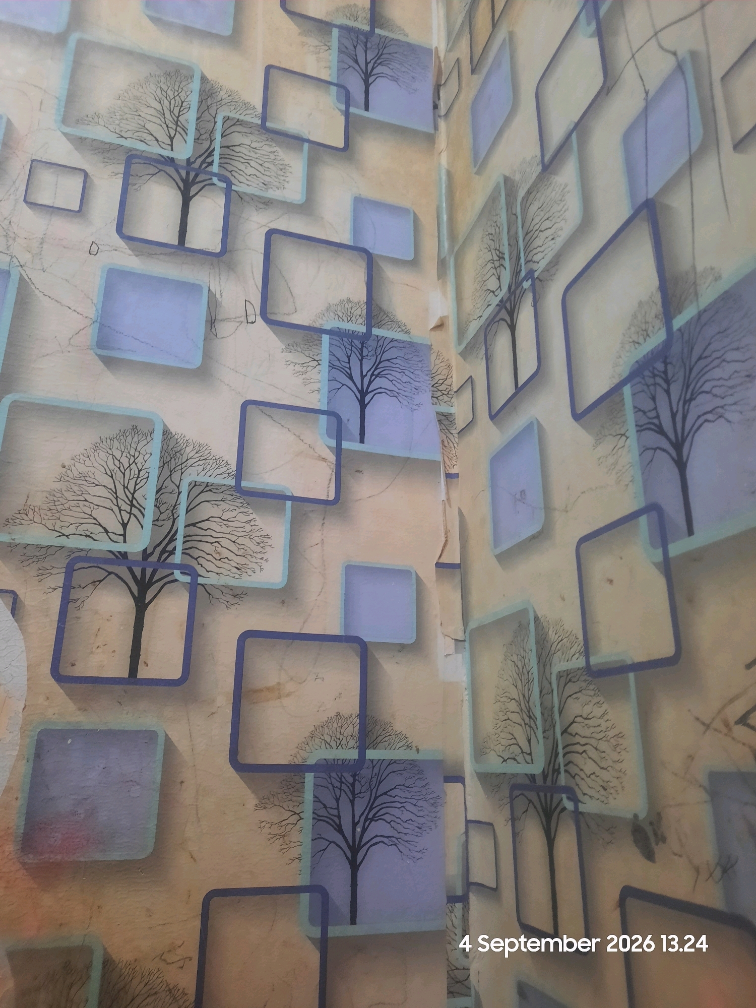 A wallpaper design features repeating rectangular frames with a faint outline of a leafless tree silhouetted within each. The frames are a gradient of light to medium blue, with some appearing translucent and others solid light blue. The background of the wallpaper is a textured, light brown color with faint grey and black scribbled lines. The wallpaper appears to be on a wall in a room, with a corner visible on the right side of the image. The lighting suggests it is indoors. The date stamp "4 September 2026 13.24" is visible in the lower right corner. The image does not provide any specific information about the location within Sukokerto, Indonesia.
