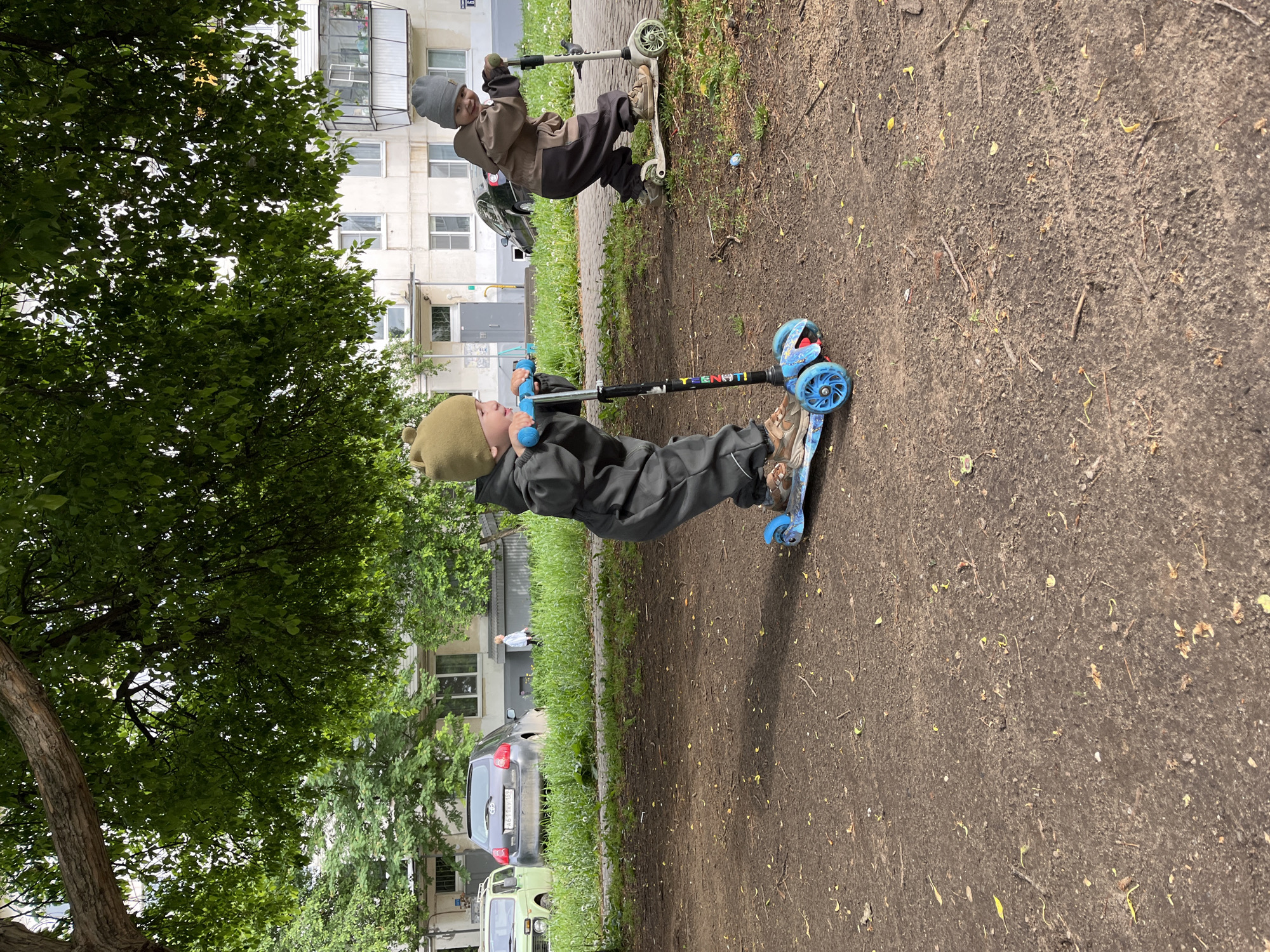 Two children are riding scooters on a dirt path next to a grassy area in Chaikovsky, Russia. One child is wearing a green hat and a gray jumpsuit, riding a blue scooter. The other child is wearing a gray hat and a brown jumpsuit, riding a white scooter. In the background are apartment buildings and cars parked along the street. A large tree partially obscures the view on the left.
