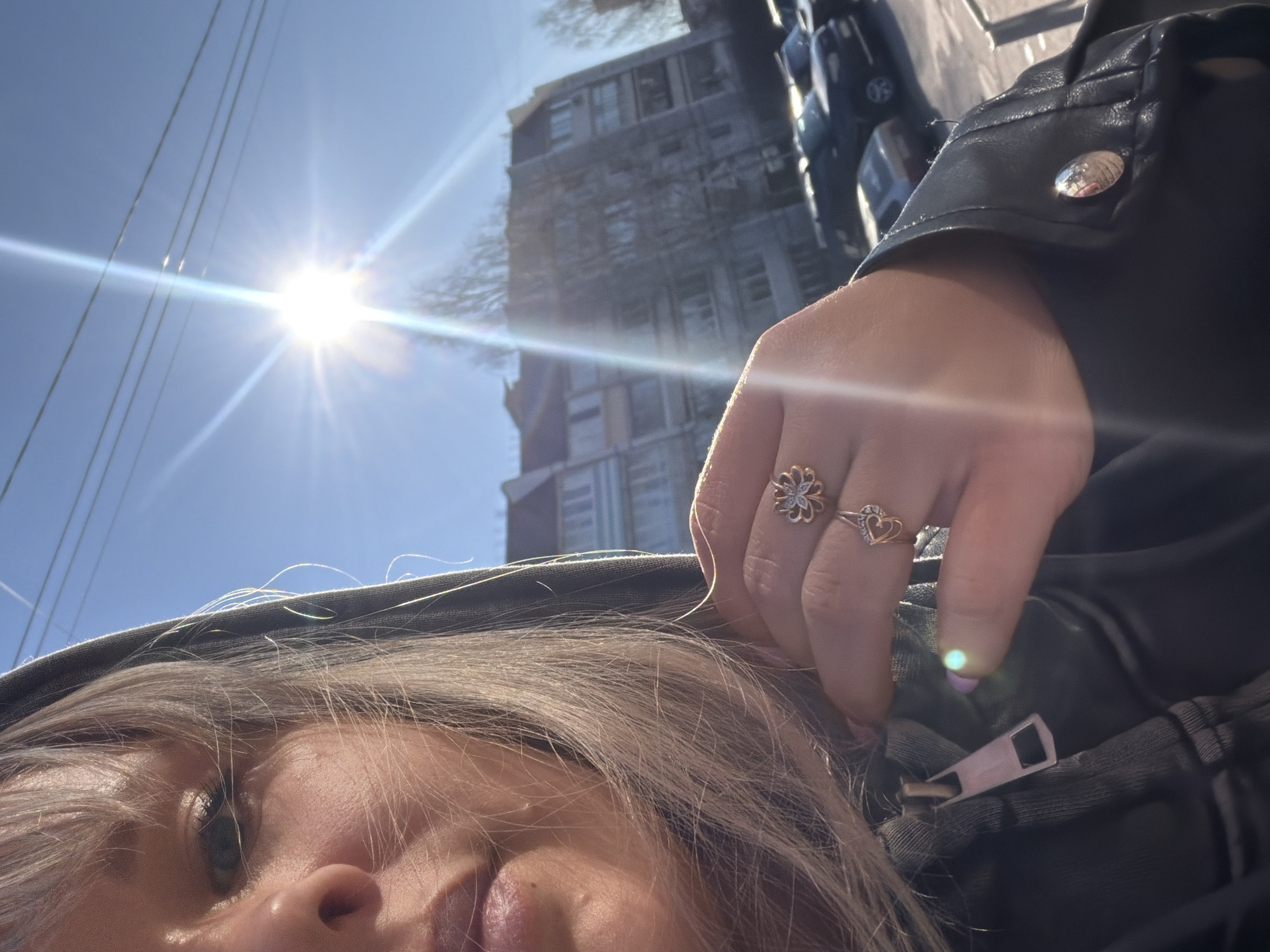 A person is looking directly into the camera, with the sun and a cityscape visible in the background. The person has light-colored hair and is wearing a dark-colored hooded jacket. Their right hand is visible, adorned with two rings: one with a floral design and another with a heart shape. The person is wearing a black leather jacket as well. A light-colored zipper pull on the jacket is visible near the person's hand. In the background, a multistory building can be seen, along with a blue sky and telephone wires. Motorbikes are parked in the background.