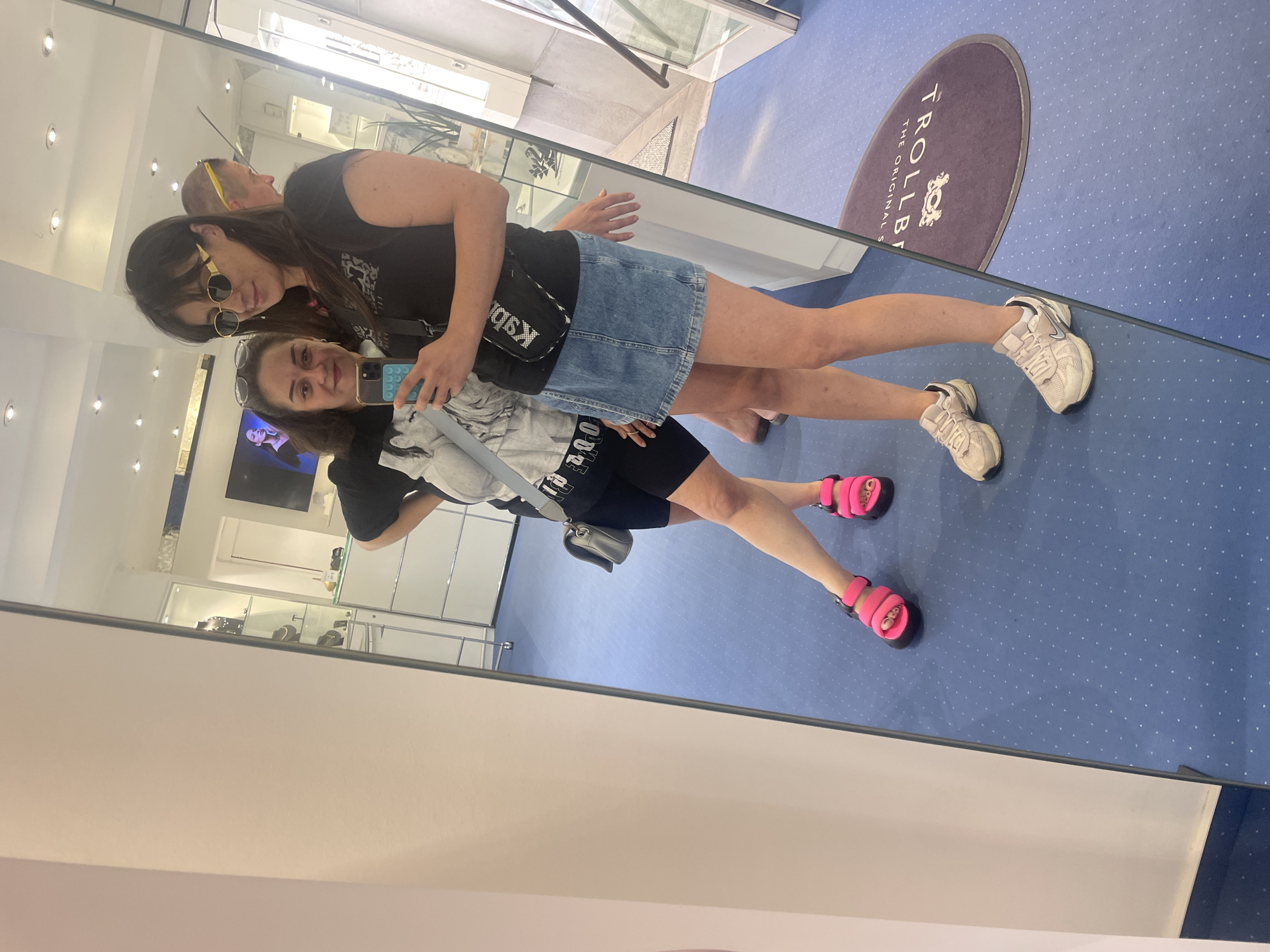 Two women are taking a mirror selfie in a retail store. The woman on the right is wearing sunglasses, a black t-shirt, and a denim skirt. She has a black fanny pack across her chest. The woman on the left is wearing a grey t-shirt with a graphic print and black shorts. She has bright pink sandals on. In the background, there are display cases filled with jewelry and other merchandise. The store has blue carpeting with a pattern of small white dots. A rug with the logo "TROLLBEADS THE ORIGINAL STORY" is visible on the floor. The presence of Trollbeads merchandise suggests the store is a retailer of this brand, which has a presence in Iserlohn, Germany. The overall environment appears to be a well-lit, modern retail space.