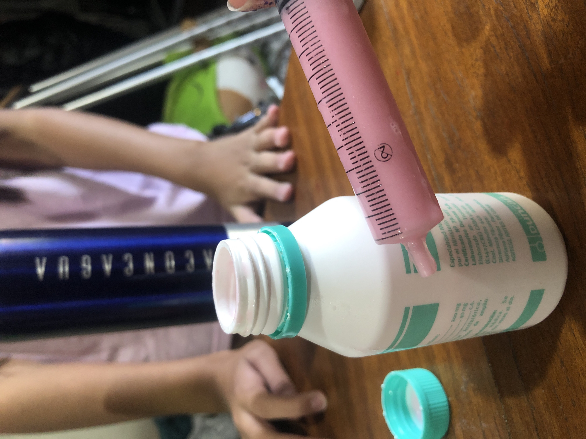 A close-up shot shows a pink liquid medication being drawn from a white bottle with a light green label and cap using a pink syringe. The bottle is partially labeled in Spanish and identifies the medication as an antibiotic from Laboratorios Duncan S.A., with the company address in Tronada (C1427CRE) in Caballito, Buenos Aires. The photograph is taken at an angle, with the focus on the medicine being extracted. A child's hand is visible near the edge of the table. An 'Aconcagua' water bottle is visible in the background.