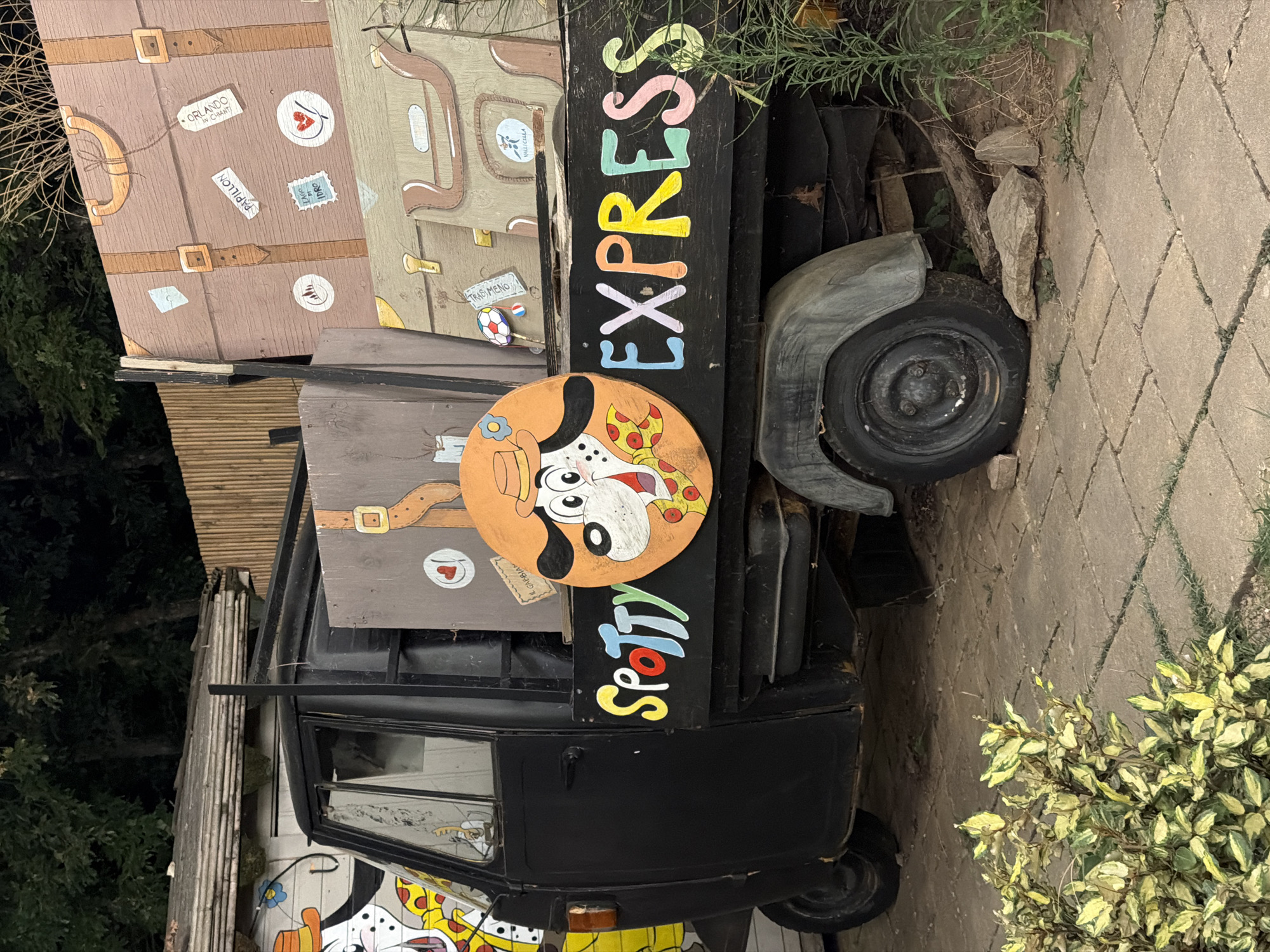 A black three-wheeled vehicle, resembling an auto-rickshaw, is parked on a paved surface. The vehicle's side panel displays colorful painted lettering spelling out "SPOTTY EXPRESS". To the left of the lettering, a circular orange panel features a cartoon drawing of a Dalmatian dog wearing a hat and a bow tie, with pizza slices near its mouth. The vehicle is loaded with large, brown, suitcase-like structures, painted with details of straps, buckles, and travel stickers. One sticker on the largest suitcase reads "ORLANDO". The background includes a wooden fence and trees, suggesting an outdoor setting. The scene is located in the city of Cavriglia, Italy, likely within an area decorated to evoke a whimsical, travel-themed atmosphere.