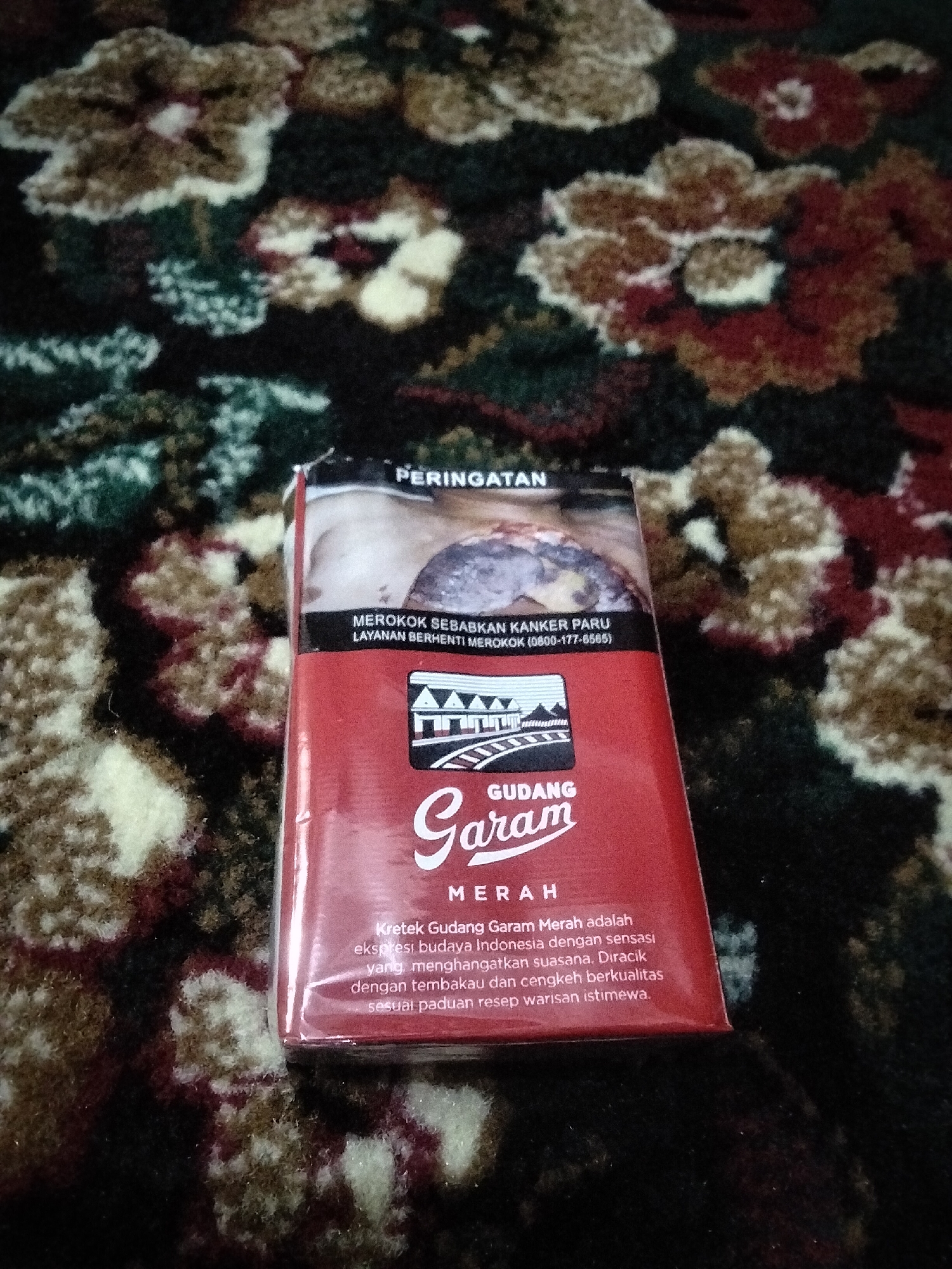 The image shows a pack of Gudang Garam Merah cigarettes on a patterned surface that appears to be a carpet or rug. The cigarette pack is red with white lettering and an image of a building. Text on the pack is in Indonesian and includes a warning about smoking causing lung cancer. There is no visible indication of the location being Lhokseumawe, Indonesia, or any people present in the image.