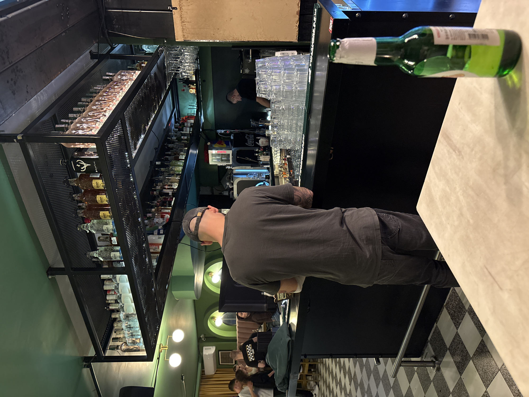 The image shows the interior of a bar in Plovdiv, Bulgaria. A customer in a gray t-shirt and baseball cap sits at the bar, which is topped with a white counter. A green beer bottle stands on the counter near the customer. Behind the counter, stacked glassware and liquor bottles are visible. Shelves stocked with various bottles extend along the wall behind the bar. A bartender is partially visible behind the bar. The bar has a black facade. Further back in the bar, other customers are seated at tables. The floor is covered in a black and white checkered pattern. Wall lamps are present on the green wall.