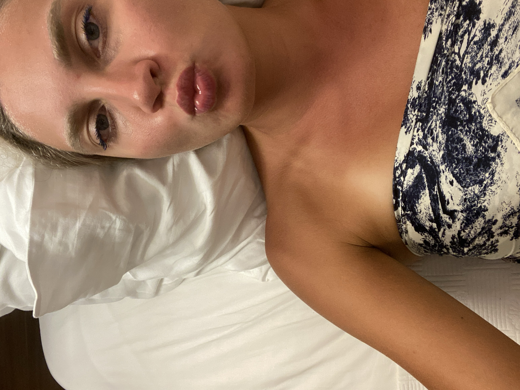A young woman is lying in bed with white sheets. She is wearing a strapless top with a blue and white abstract pattern. Her lips are pursed, and she is wearing blue eyeliner. The lighting suggests it is daytime. There is no information about the city of Aksu, Turkey in this image.