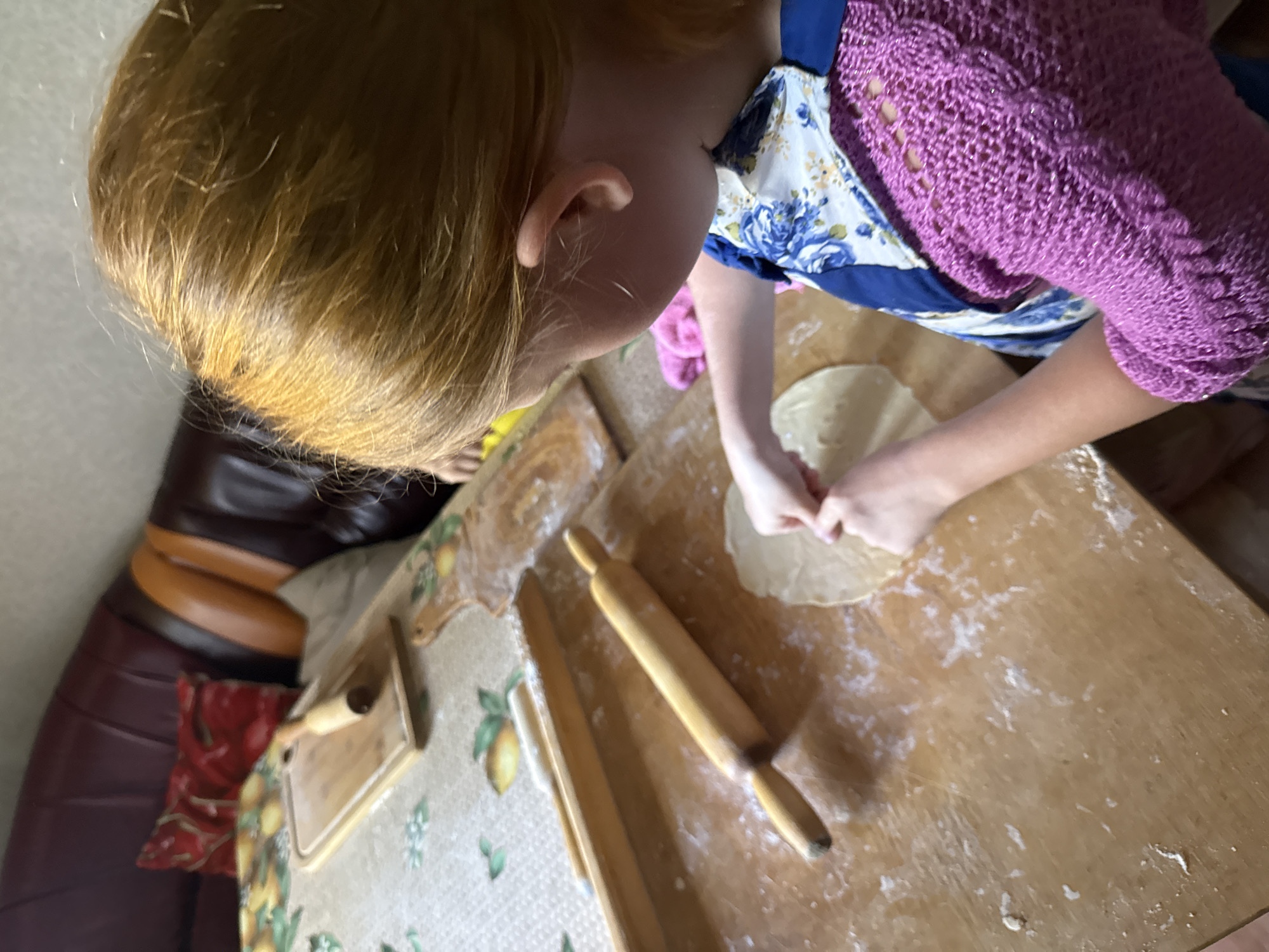 A young girl with blonde hair is shown from behind, as she prepares dough on a wooden surface. She is wearing a blue and white floral apron over a pink knitted sweater. A rolling pin is visible on the table in front of her, along with a flattened piece of dough. In the background, a portion of a brown leather couch with red and yellow accents can be seen. The overall setting appears to be a domestic kitchen or dining area, possibly in Valday, Russia, given the context.