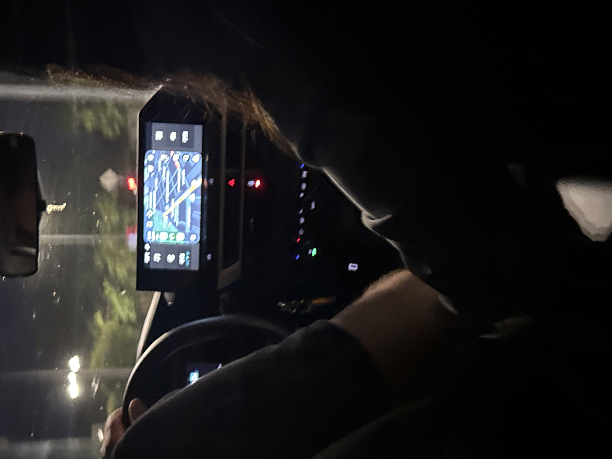 The image depicts the interior of a car at night, likely in Burgas, Bulgaria, based on the context provided by the prompt, though this cannot be definitively confirmed from the image alone. A person's hands are visible on the steering wheel, suggesting they are driving. Another person's arm and shoulder are visible in the foreground. The car's infotainment screen is displaying a navigation map. The environment outside the car appears to be dark with some streetlights visible. The overall mood is intimate and possibly romantic, given the way the hands are positioned on the steering wheel, though this is subjective.