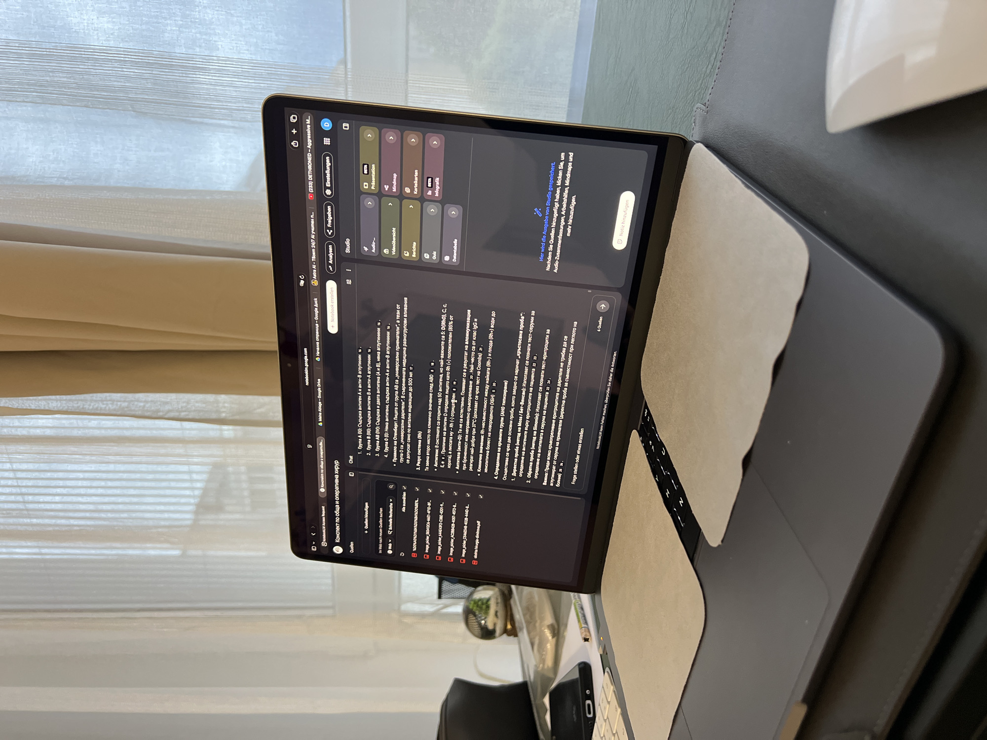 The image shows a laptop open on a desk. The laptop screen displays a web page with text in Bulgarian, likely a document or presentation. There are several tabs open in the browser, including one labeled "Google Duck" and another with a question mark icon. The laptop is a dark color and appears to have a keyboard and trackpad. On the desk, next to the laptop, is a dark smartphone. The desk is covered with a dark gray surface, and there is a white computer mouse visible on the right side of the frame. In the background, a sheer white curtain is drawn across a window, allowing diffused natural light to enter the room. The overall impression is that of a workspace, possibly a home office. There is no visual information in the image that allows for the precise identification of the location within the city of Burgas, Bulgaria.