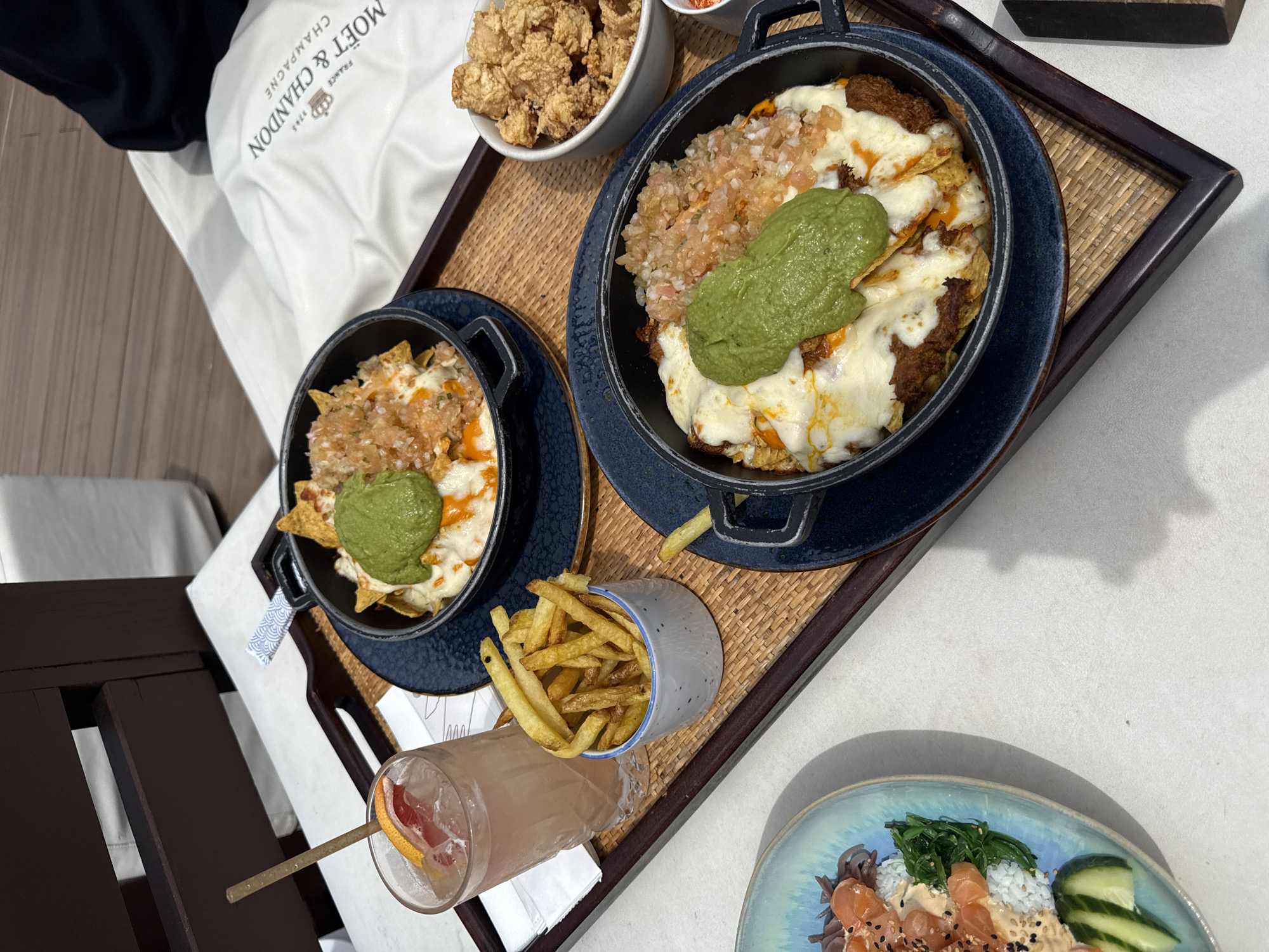 The image captures a high-angle, close-up view of a dining spread arranged on a light-colored tabletop, likely in an outdoor or semi-outdoor setting in Barcelona, Spain, as indicated by the bright, directional sunlight casting distinct shadows.
The primary elements are presented on a rectangular woven rattan tray with dark wooden borders. On the left side of the tray, a textured, clear glass contains a pink beverage with ice, garnished with a slice of grapefruit on the rim and a beige straw inserted. Immediately to its right, a small white ceramic cup with blue specks holds a portion of golden-brown french fries.
Two identical black cast iron-style mini-skillets are centered on the tray, each resting on a dark blue, patterned ceramic plate. Both skillets contain a dish composed of a base (likely nachos or loaded fries) generously covered with melted white cheese, a scoop of green guacamole, and a topping of finely diced red and white salsa. Traces of an orange sauce are visible beneath the cheese.
To the right of the lower skillet, a small white bowl contains multiple irregularly shaped, golden-brown fried pieces, consistent with popcorn chicken or similar fried bites.
In the lower left corner, partially visible on a light blue, irregularly shaped plate off the main tray, is a portion of a poke bowl. Observable components include white rice, green seaweed, slices of cucumber, cubes of salmon, sesame seeds, and a creamy, light-colored sauce.
In the upper right background, a white cushion or pillow is visible, displaying the inverted black text "MOËT & CHANDON CHAMPAGNE FRANCE" along with a crown emblem. Dark brown wooden elements, possibly part of a chair or table, are visible in the upper left background. No individuals are present within the frame.