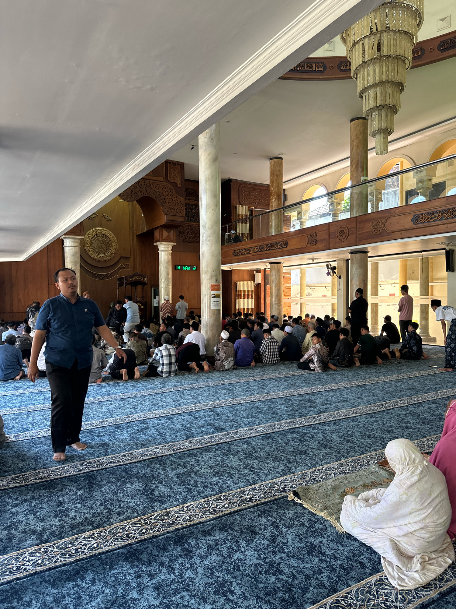 This image depicts the interior of a mosque, likely in Malang, Indonesia, during a prayer gathering. The space is filled with people, predominantly men, seated or kneeling on a blue carpet with intricate white patterned borders. The carpet is arranged in rows, suggesting an organized prayer service.
In the foreground, a man in a blue shirt and black pants walks across the carpet, facing away from the camera. His stance suggests he is participating in or moving within the prayer space. Behind him, a large congregation is visible, with individuals in various attire, many wearing traditional Islamic clothing such as peci (caps) and sarongs. Some are facing the front of the mosque, presumably towards the Qibla.
The mosque interior features tall, round columns and wooden architectural elements, including decorative carvings and what appears to be a mezzanine level with glass railings. A large, multi-tiered chandelier hangs from the ceiling, illuminating the space. The walls are adorned with intricate patterns and calligraphy, typical of Islamic mosque architecture. Natural light enters through large arched windows on the upper level, providing additional illumination. The overall atmosphere appears to be one of reverence and communal worship.
