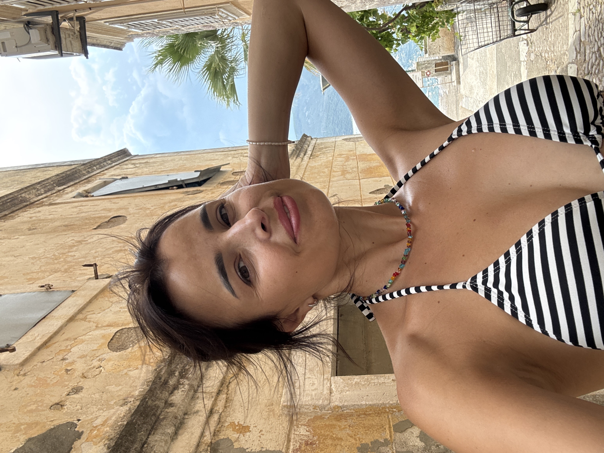 A woman with dark hair pulled back is taking a selfie in an alleyway. She is wearing a black and white striped bikini and a colorful beaded necklace. She has one arm behind her head, and she is smiling at the camera. The background shows a weathered yellow building on the left and a similar building on the right, with a palm tree and blue sky visible between them. In the distance, mountains and the sea can be seen. A shopping cart is visible on the right side of the image.