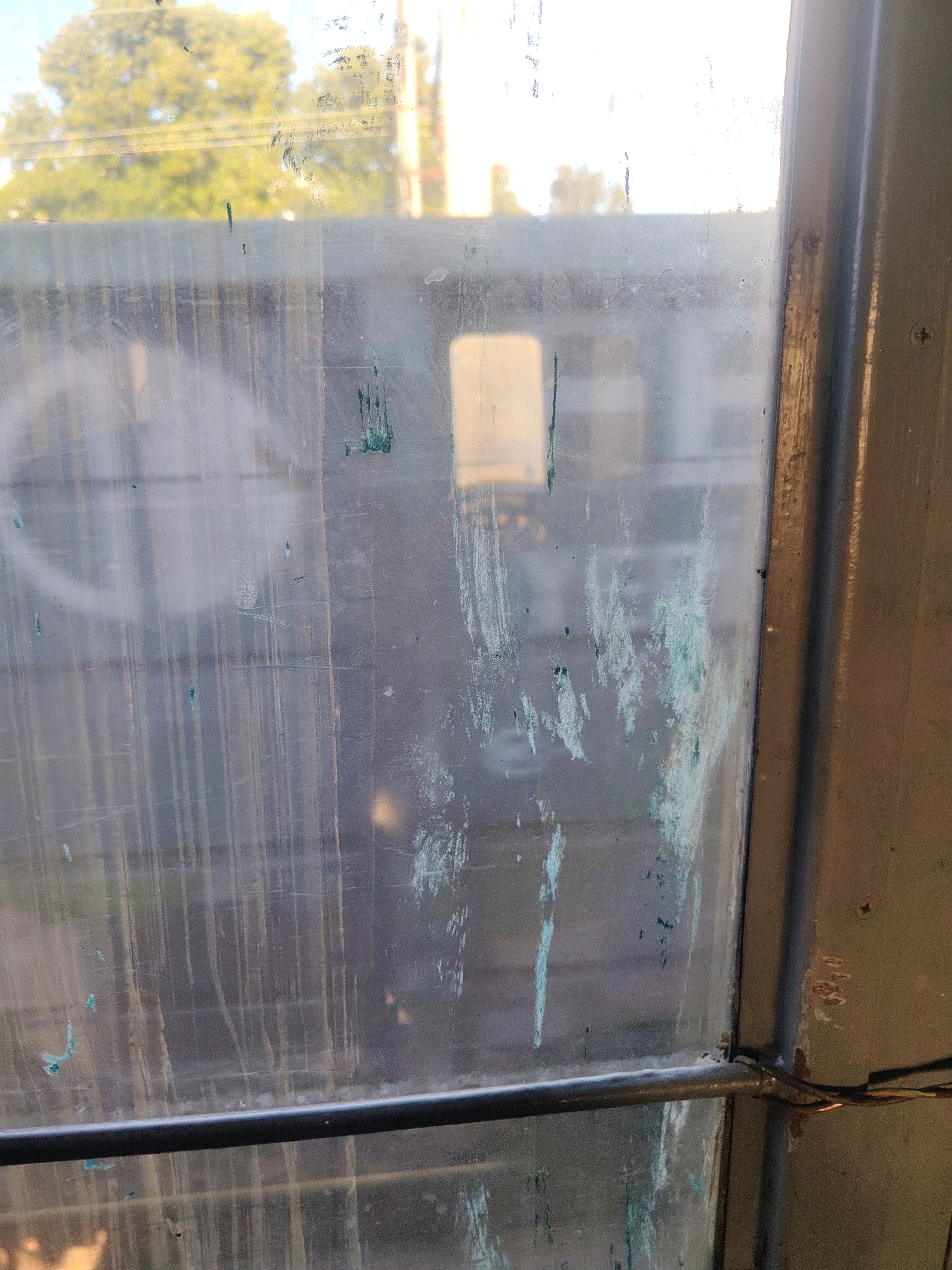 The image depicts a close-up view of a dirty window pane with some green streaks of paint or residue on it. Through the window, a blurry outdoor scene is visible, featuring trees and possibly some buildings. The lighting suggests it might be daytime, with sunlight filtering through. The window frame appears to be made of metal and wood, showing signs of wear and tear. The overall impression is one of neglect or disrepair. There is no discernible activity or event taking place. No text is visible in the image. The location context provided is Kyiv, Ukraine.