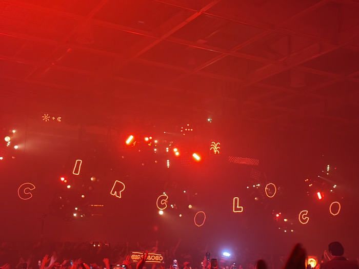 The image depicts an indoor electronic music event, specifically a Circoloco party, held at the Paris La Défense Arena in Nanterre, Paris, France. The scene is dominated by intense red stage lighting that permeates the entire space, creating a hazy atmosphere. Above a large crowd of attendees, the word "CIRCÓLOGO" is spelled out in suspended, glowing orange-red outline letters. Below these letters, a screen or banner displays "CIRCOLOCO" in a red text. Various bright stage lights are visible, some projecting outwards, others appearing as point sources. Illuminated symbols including a sun, a plus sign, a crescent moon, and a stylized plant or tree are visible amongst the lighting fixtures. In the lower foreground, a dense congregation of people with raised hands, many holding illuminated mobile phones, indicates active engagement with the performance. The high ceiling of the venue is partially visible in the upper background, obscured by the red light and haze.