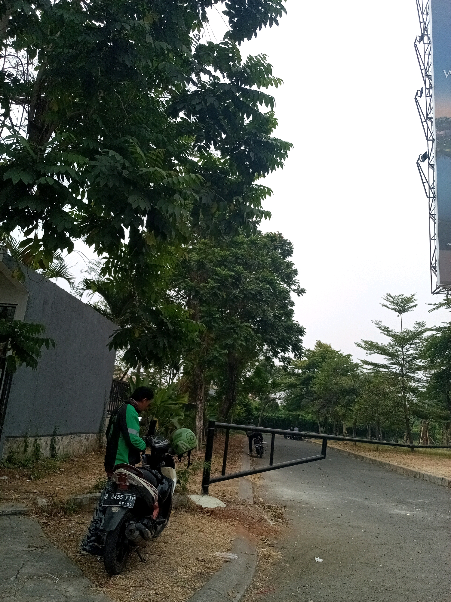 The image depicts an outdoor scene in Bekasi, Indonesia, featuring a person standing next to a parked motorcycle. The individual, wearing a green and black jacket, is looking down at the motorcycle's handlebars, possibly interacting with a device attached there. A green helmet with a white decal is hanging on the motorcycle's rear. The motorcycle, a black scooter, has a license plate that reads "B 3455 FIP 07-27". The immediate surroundings include a grey building on the left, lush green trees, and a paved road with a black barrier gate. The ground is covered in dry leaves and dirt. In the background, more trees and a billboard are visible under an overcast sky.