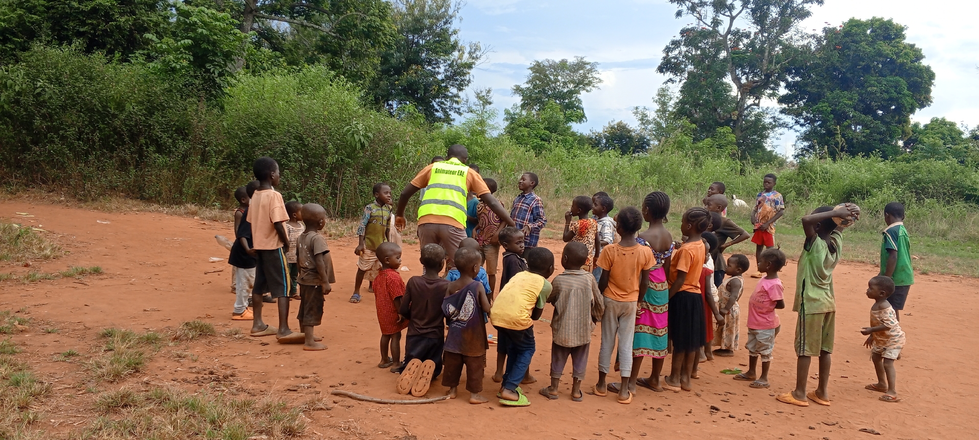 In Ngalula, Democratic Republic of the Congo, a group of approximately 25 children of varying ages stand on reddish-brown earth in an outdoor setting. They are gathered around a man who is wearing a yellow safety vest, which has "Animateur EAE" printed on it. The children are dressed in an assortment of casual clothing, including shirts, dresses, and shorts of various colors and patterns. The environment includes trees and bushes in the background, indicating a somewhat rural or undeveloped area. The sky is partly cloudy. Some of the children are looking at the man, while others are looking elsewhere, seemingly engaged or curious. The ground is uneven with some patches of grass and a stick is laying on the ground near a pair of shoes.