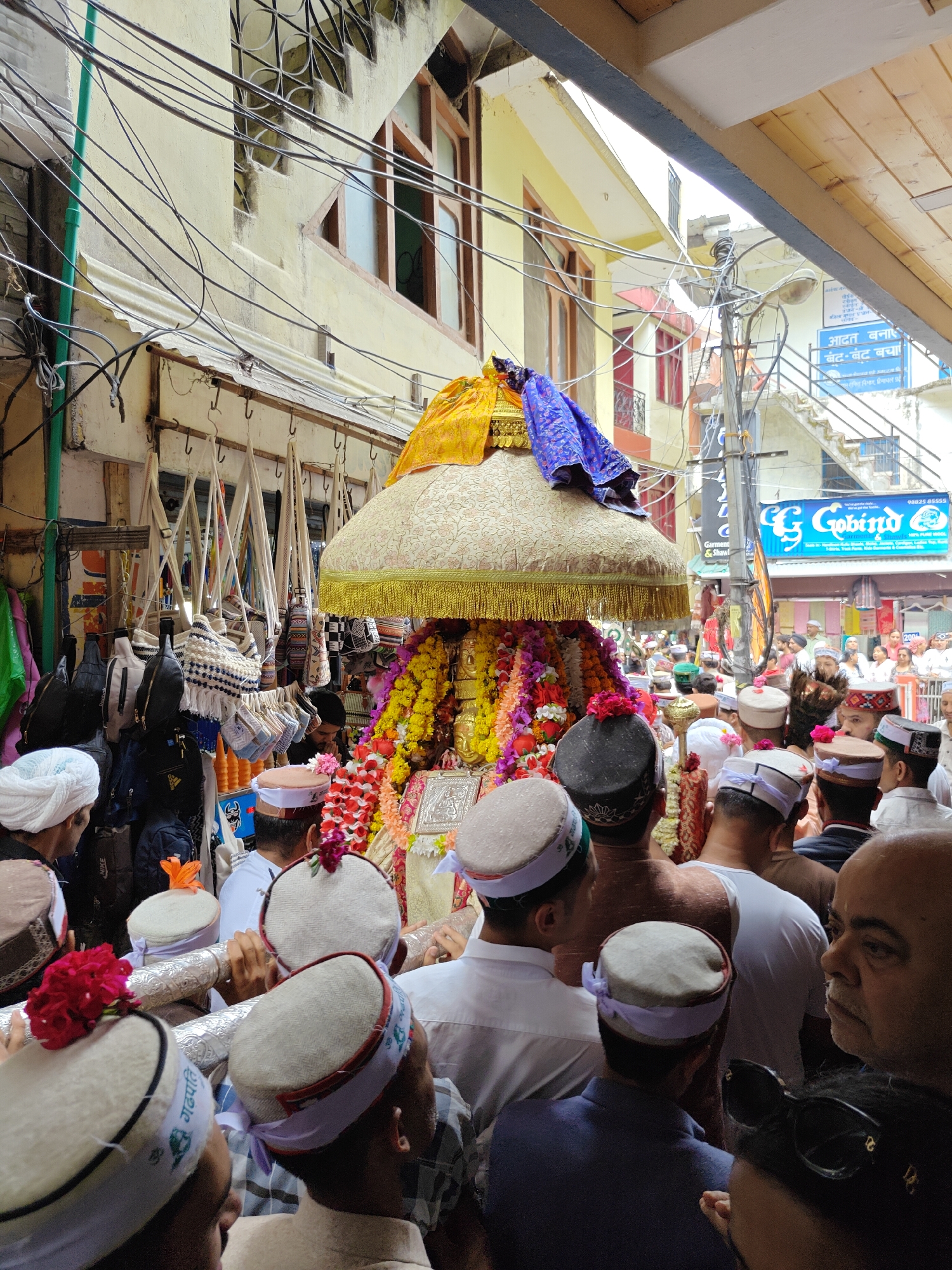 A vibrant religious procession unfolds on a crowded, narrow street in Kasol, India. The central focus is a highly decorated palanquin, or rath, carrying a gilded idol or deity figure. The palanquin is topped with a large, ornate, gold-embroidered canopy fringed with gold tassels, and draped with purple fabric. It is profusely adorned with garlands of bright yellow, orange, and red flowers.
Numerous people, predominantly men, are gathered around the palanquin, some of whom are actively carrying it on their shoulders. Many in the crowd wear distinctive traditional Himachali caps, which are typically white or cream-colored with patterned bands, some featuring small red flowers as decoration. The atmosphere appears reverent and celebratory.
The scene is set amidst old, multi-story buildings with traditional wooden-framed windows and balconies. Numerous black electrical wires crisscross erratically overhead, characteristic of Indian towns. To the left, a shop stall displays various hanging bags and pouches, suggesting a market area.
Visible text on a blue signboard in the background includes Hindi words: "गोविंद" (Govind) and "गारमेंट्स & शोए" (Garments & Shoes), along with a phone number, "9824 45555". Another partially visible blue signboard shows Hindi text like "आदत बनाइ" (Make a habit) and "बैट-बैट बचा" (Save bit by bit, or possibly referring to 'battery'). On some of the caps, Hindi script such as "गढपति" (Gadpati) and the 'Om' symbol are discernible. The time of day is daytime, with natural light illuminating the bustling street.