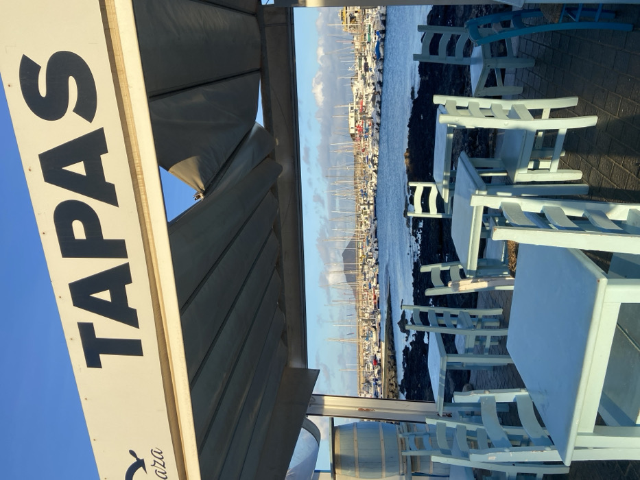The image is taken from inside a restaurant in Corralejo, Spain, specifically at the coordinates 28.741 latitude and -13.866 longitude. The restaurant's awning, which is made of canvas material, is partially open and shows the harbor with boats and a distant island, while the blue sky is visible in the upper left corner of the image. The awning is marked with the word "TAPAS" in large, bold, black letters. The image focuses on the restaurant tables and chairs which are made of light blue wood. They are positioned on a black sand floor, with a view of the harbor. There are empty tables and chairs, suggesting that no one is currently using the seating area.