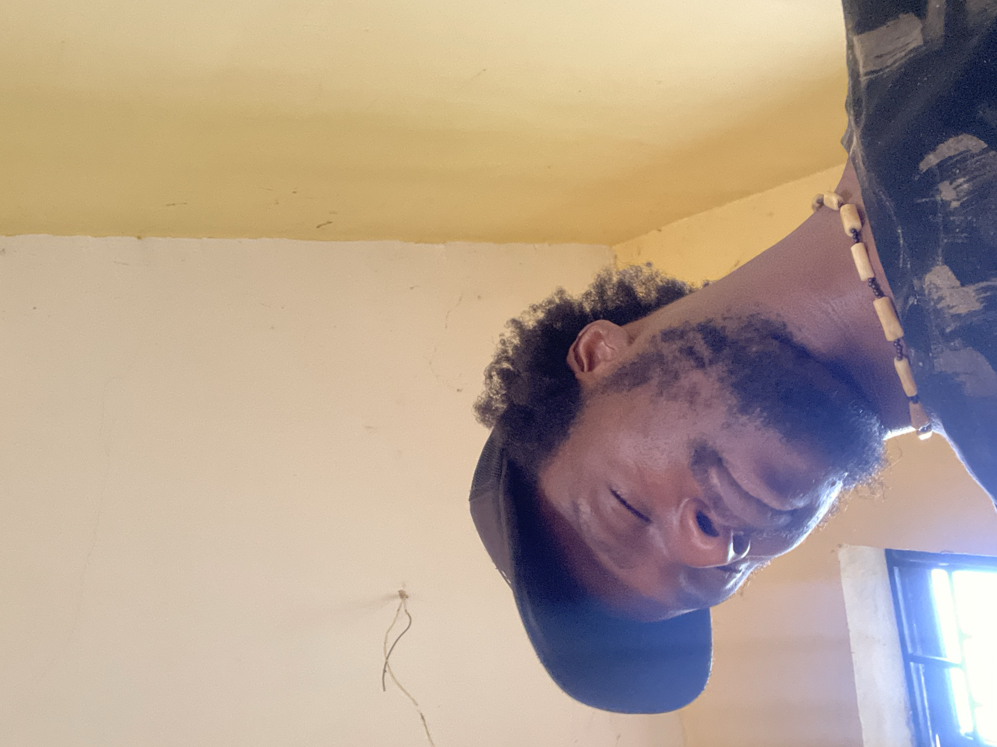 A close-up, low-angle selfie taken indoors shows a Black male wearing a black baseball cap, a black t-shirt with a distressed print, and a beaded necklace. His eyes are closed, and his head is tilted slightly. The background consists of a pale cream ceiling with a thin crack and a light yellow wall. A window with horizontal bars is visible in the lower left corner, letting in bright light. The setting appears to be a residential interior in Abuja, Nigeria.