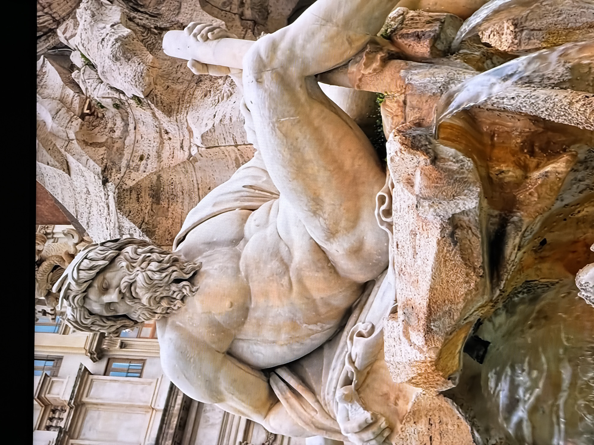 The image shows a close-up of a monumental stone sculpture of a reclining male figure with a beard and laurel wreath. The sculpture is part of a fountain, with water cascading over rough-hewn rocks. The figure's musculature is detailed, and he is draped in fabric. In the background, architectural elements of a building with arched windows are visible. The lighting suggests it is daytime, and the overall scene is grand and artistic. The prompt's request to place this in Chirchik, Uzbekistan, is noted, but the visual evidence strongly suggests a European location, likely Rome, given the style of architecture and sculpture, which are characteristic of Italian Baroque art and fountains. Without further information or visual cues from the image itself, it is not possible to confirm or deny the Chirchik, Uzbekistan location.