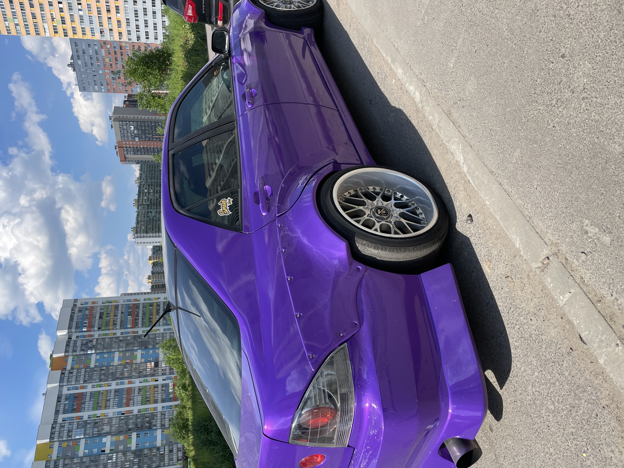 The image displays a customized deep metallic purple sedan, identifiable as a Lexus IS (XE10 generation), parked on an asphalt road in a bustling urban residential area under a bright, sunny sky with scattered white clouds. The vehicle features riveted wide-body fender flares over the rear wheels, multi-spoke chrome or polished silver deep-dish wheels with low-profile tires, and a large, black single exhaust tip exiting from the driver's side of the custom-painted rear bumper. Aftermarket taillights with clear/smoked lenses and red circular elements are installed. A black antenna is visible on the roof, and a "Smoke Shop" sticker is affixed to the rear passenger window.
In the background, multiple tall residential buildings dominate the skyline. The most prominent structure, situated directly behind the car, is a high-rise characterized by a white facade and numerous colorful window panels in shades of blue, yellow, green, orange, and grey. Other residential high-rises, varying in height and color (some predominantly grey, others with orange and yellow accents), extend into the distance. Lush green trees and dense foliage are present between the road and the buildings. To the right of the purple sedan, the rear section of a dark-colored SUV, consistent with a Kia Sportage, is partially visible. The immediate foreground shows an asphalt road bordered by a concrete curb, with strong shadows cast by the vehicles and environment, indicating intense overhead sunlight.