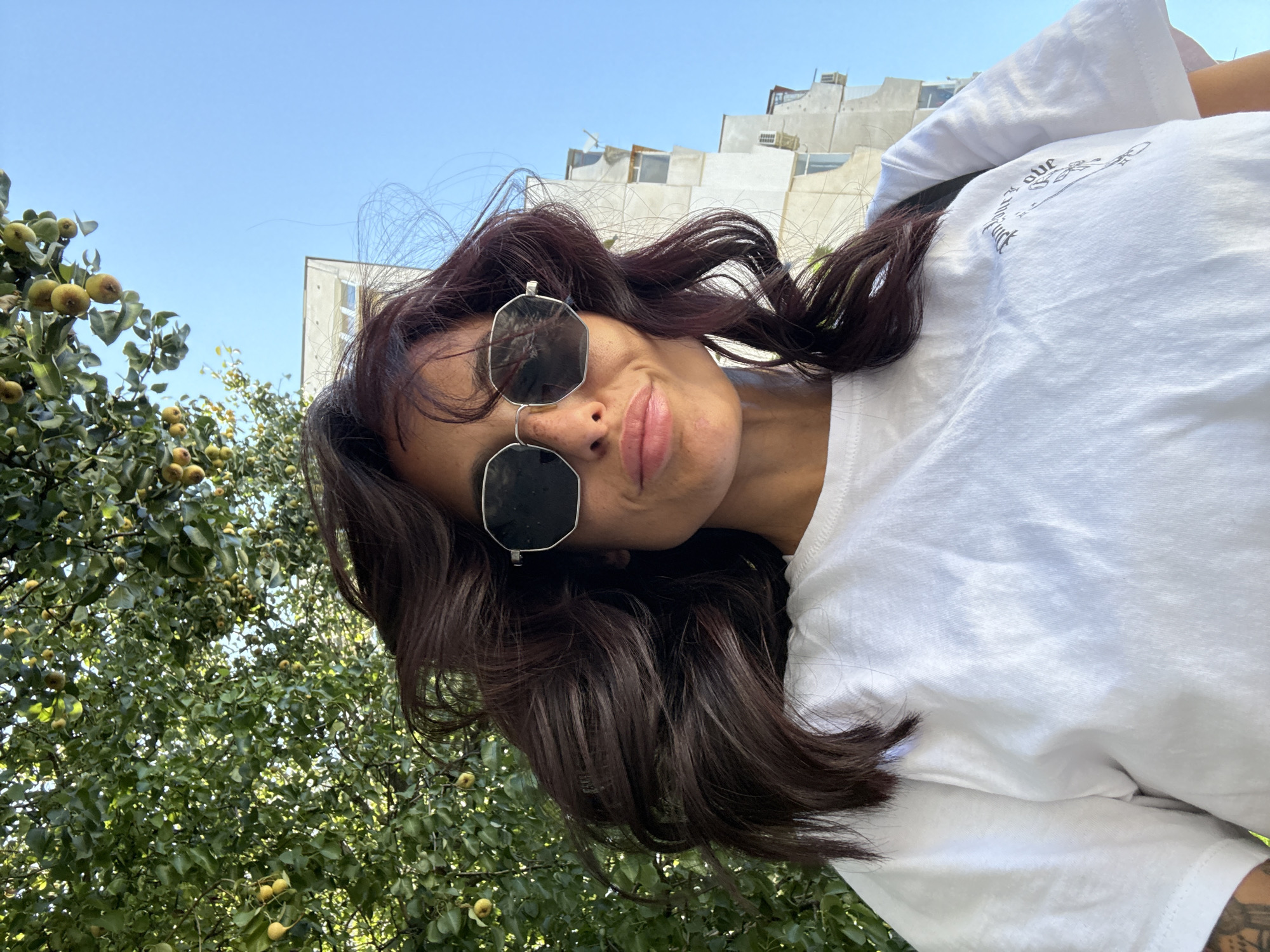 A young woman with dark, wavy hair, wearing sunglasses and a white t-shirt, is standing outdoors. She appears to be looking at the camera. Behind her, a tree with small, light-colored fruits is visible, along with a light-colored building and a clear blue sky. The scene suggests a daytime setting, possibly in a residential area. The image was taken in Pleven, Bulgaria.