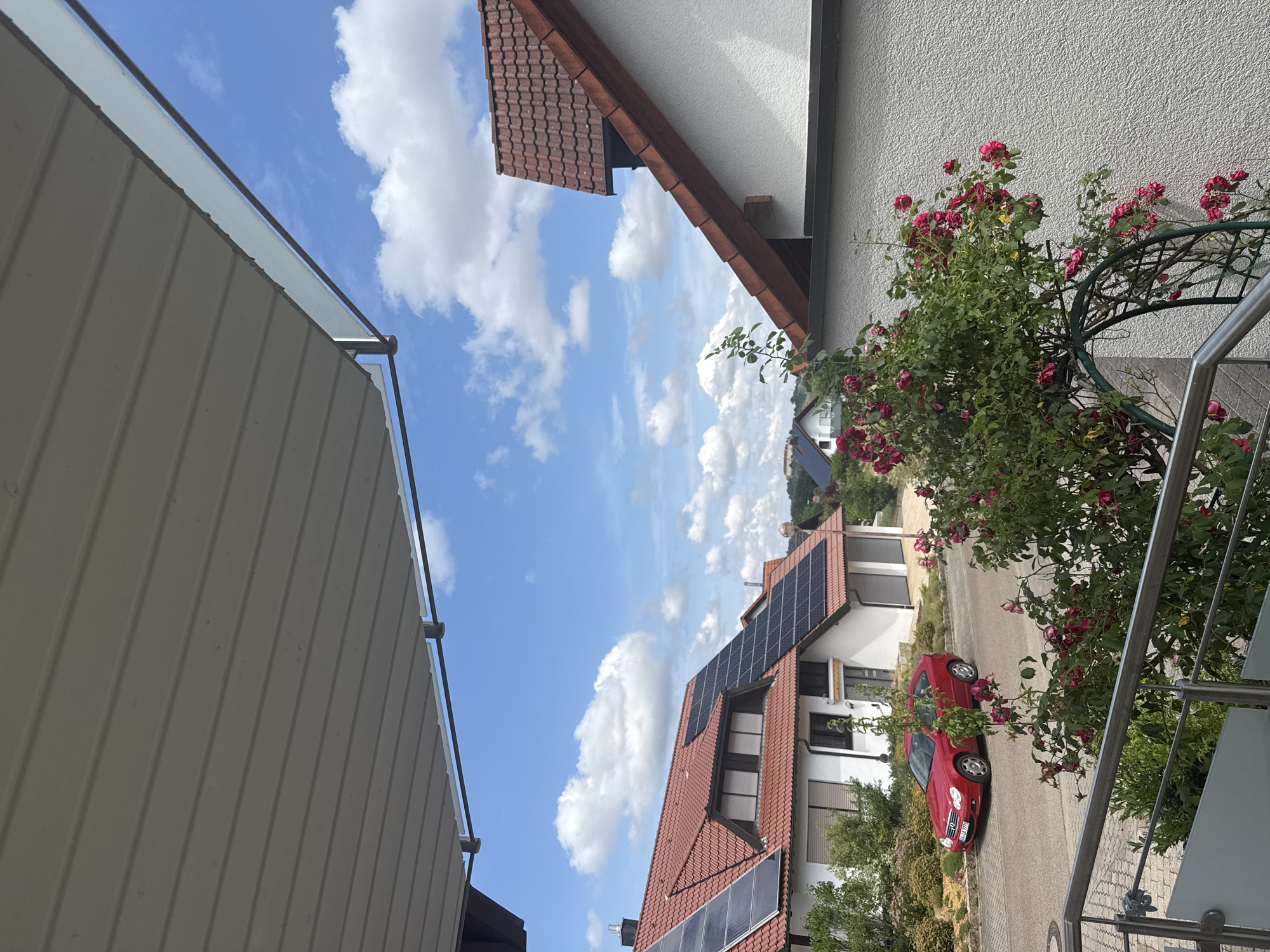 The image depicts a residential street scene on a bright, sunny day with scattered clouds in a blue sky. In the foreground, a white textured wall and a metal railing are visible, with a rose bush in bloom arching over a metal trellis. Beyond the wall, a red car is parked on the left side of the street in front of a white house with a red tiled roof and solar panels. Another house with a red tiled roof and solar panels is visible to the right, partially obscured by the foreground wall. The environment suggests a suburban or rural setting in the city of Unknown, Unknown.