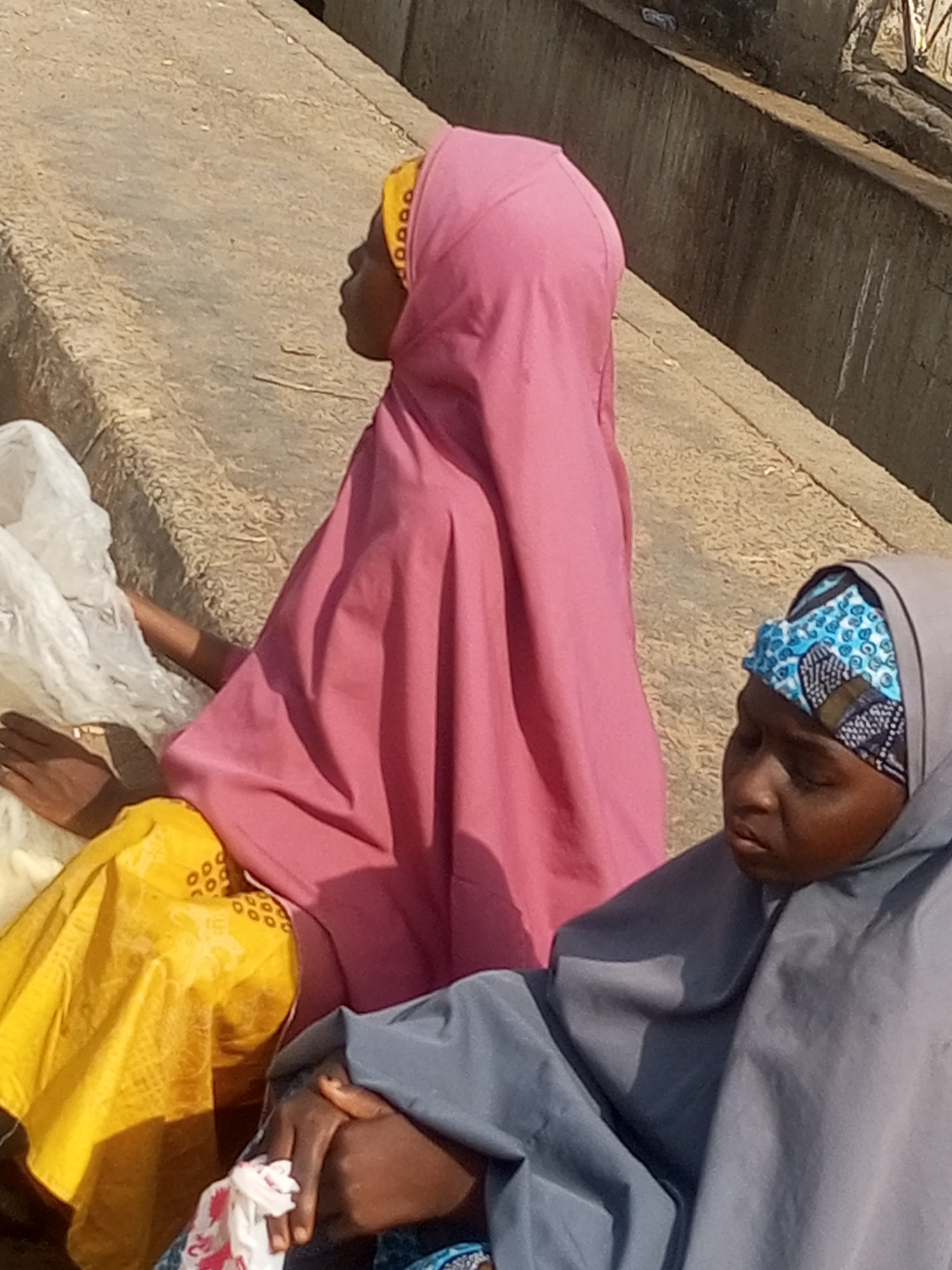 In Tempure, Nigeria, two individuals are seated on a textured, sunlit surface, possibly a raised platform or steps, adjacent to a concrete wall. The person on the left is covered in a bright pink hijab and a yellow garment, with their hands resting on a white plastic bag. To their right, another person wears a grey hijab and a blue and white patterned headscarf. Their head is bowed, and their hands are clasped, with a small, patterned cloth bundle held between them. The background is dominated by the rough texture of the ground and the grey concrete wall.