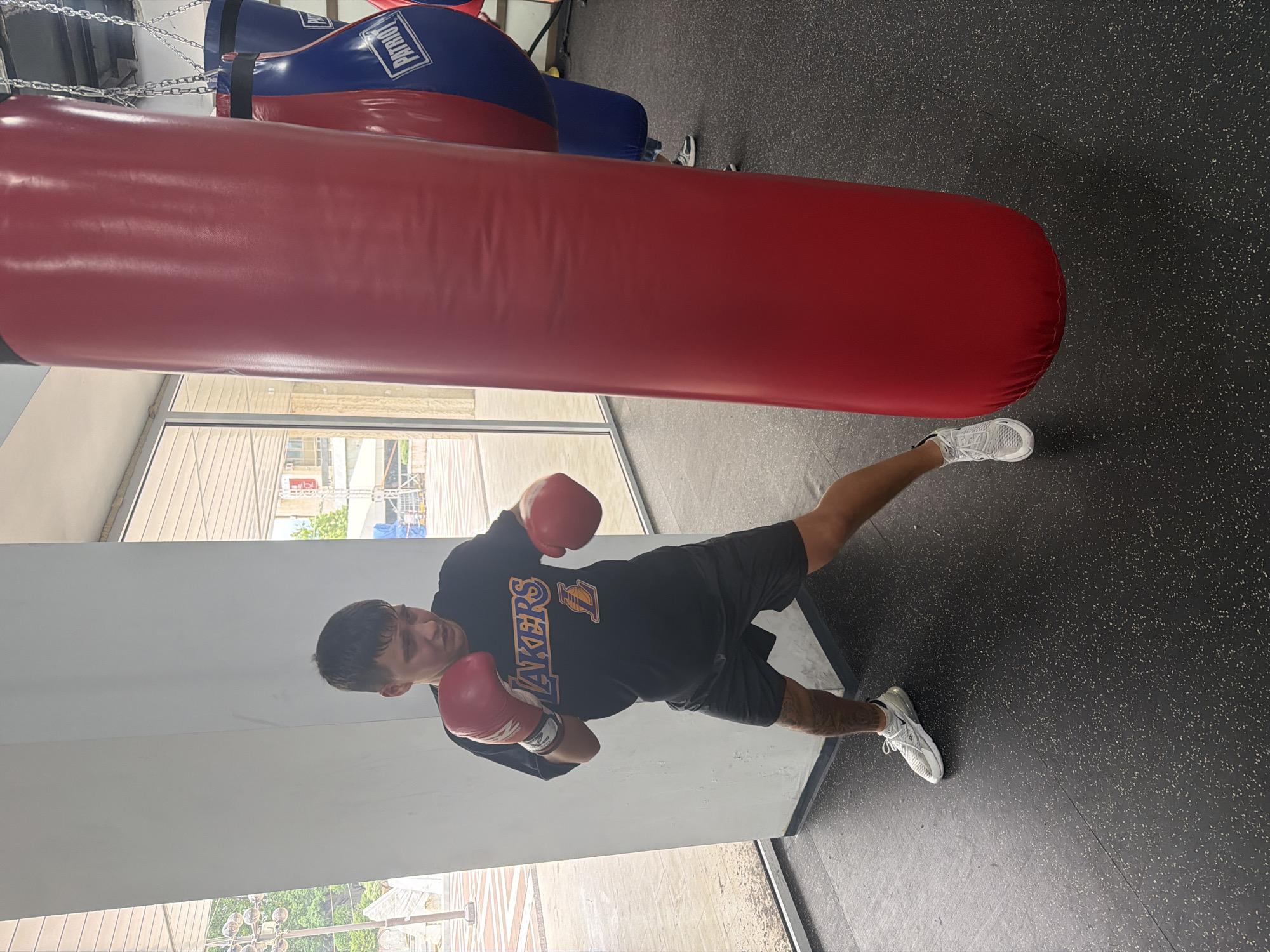 A man wearing a black t-shirt with "Lakers" printed on it, black shorts, white sneakers, and red boxing gloves is posing in a boxing stance in front of a large red punching bag in a gym. There is a blue and red punching bag in the background. The gym has a black floor with white speckles, white walls, and large windows. Outside the windows, buildings and foliage can be seen. The location of the gym is in a city. The person is not in Blagoevgrad, Bulgaria.