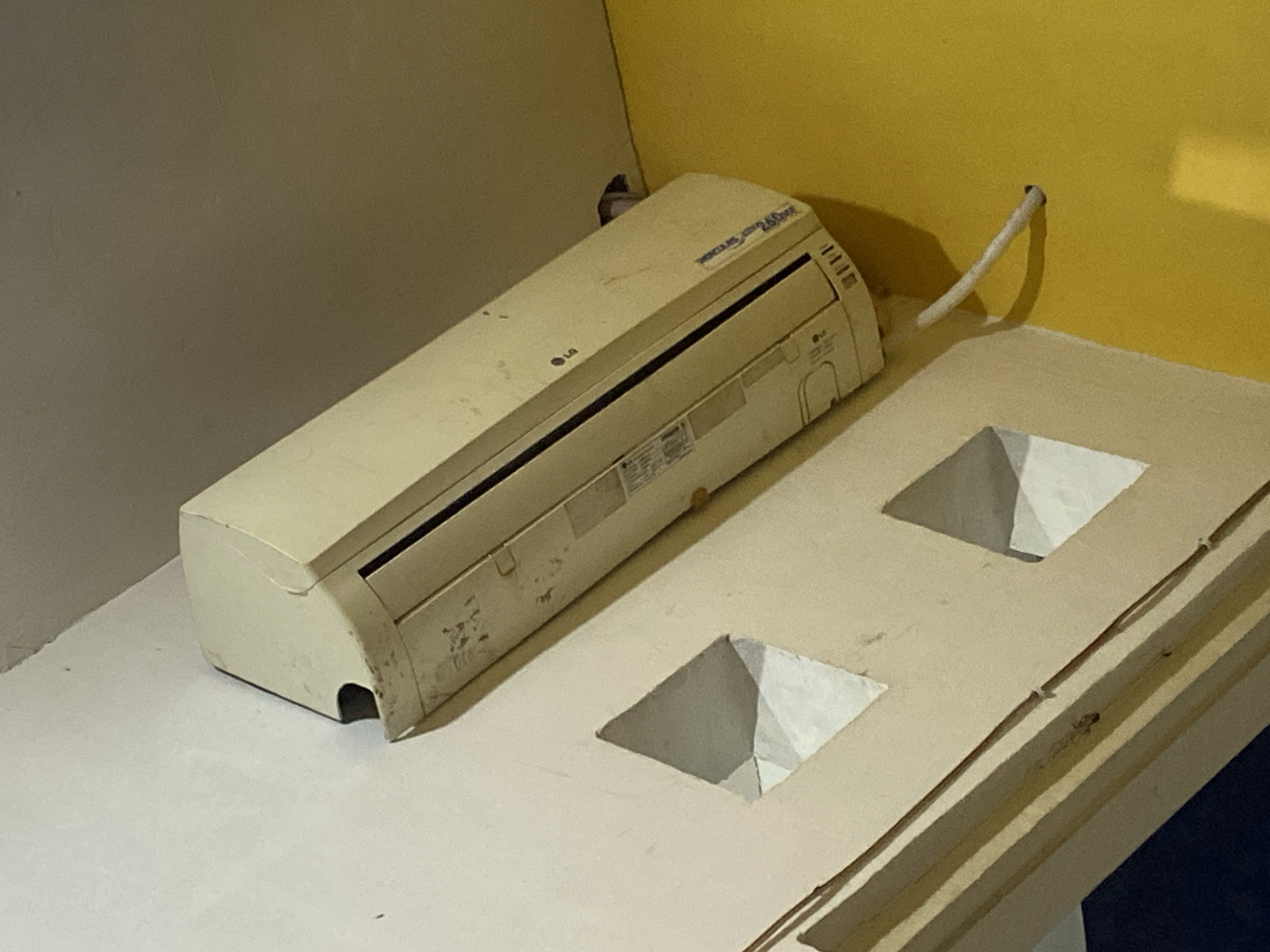 The image shows an indoor scene featuring an off-white LG air conditioning unit mounted on a textured, light-colored wall. The air conditioner is positioned at an angle, with its right side closer to the camera. To the left of the air conditioner, the wall has decorative diamond-shaped openings. The ceiling above appears to be a muted yellow or beige color, and the wall to the right is a bright yellow. A white pipe extends from the air conditioner and disappears into a small hole in the yellow wall. There is no indication of people or specific activities in the image. The lighting suggests an indoor environment, possibly a room or hallway. The location is identified as Gendingan, Indonesia.