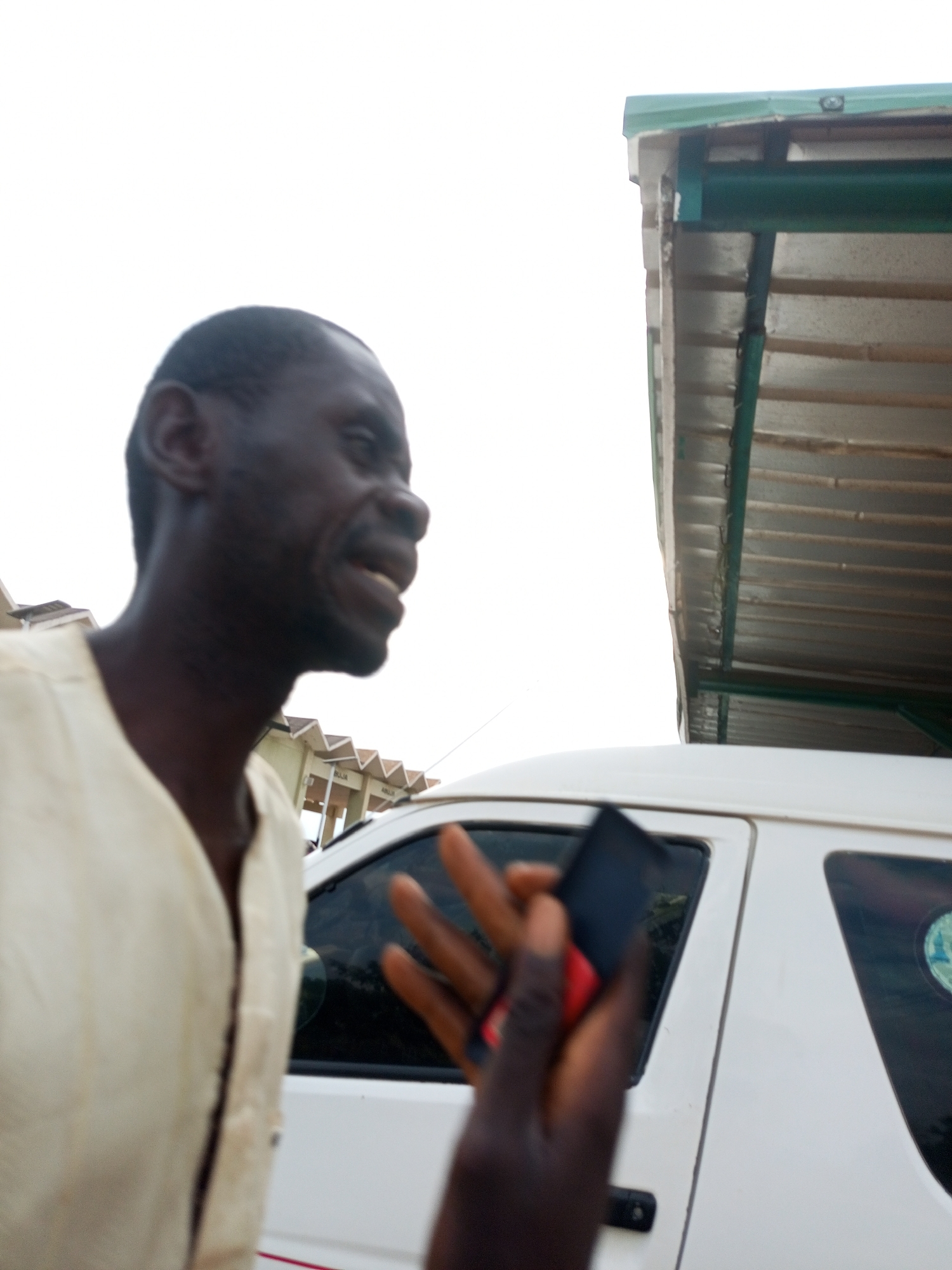 An outdoor, medium shot captures a dark-skinned man in profile, wearing a light-colored, collared shirt. He appears to be speaking, with his mouth open. His left hand is raised, holding a dark flip phone with a red button. Behind him, a white vehicle, possibly a van, is visible. The vehicle has tinted windows and a portion of its roof is obscured by a corrugated metal structure with a green trim. The background is a bright, overcast sky. The scene suggests an informal setting, possibly a roadside or a marketplace, given the presence of the vehicle and the makeshift shelter. The overall impression is a candid moment, possibly a conversation or a transaction.